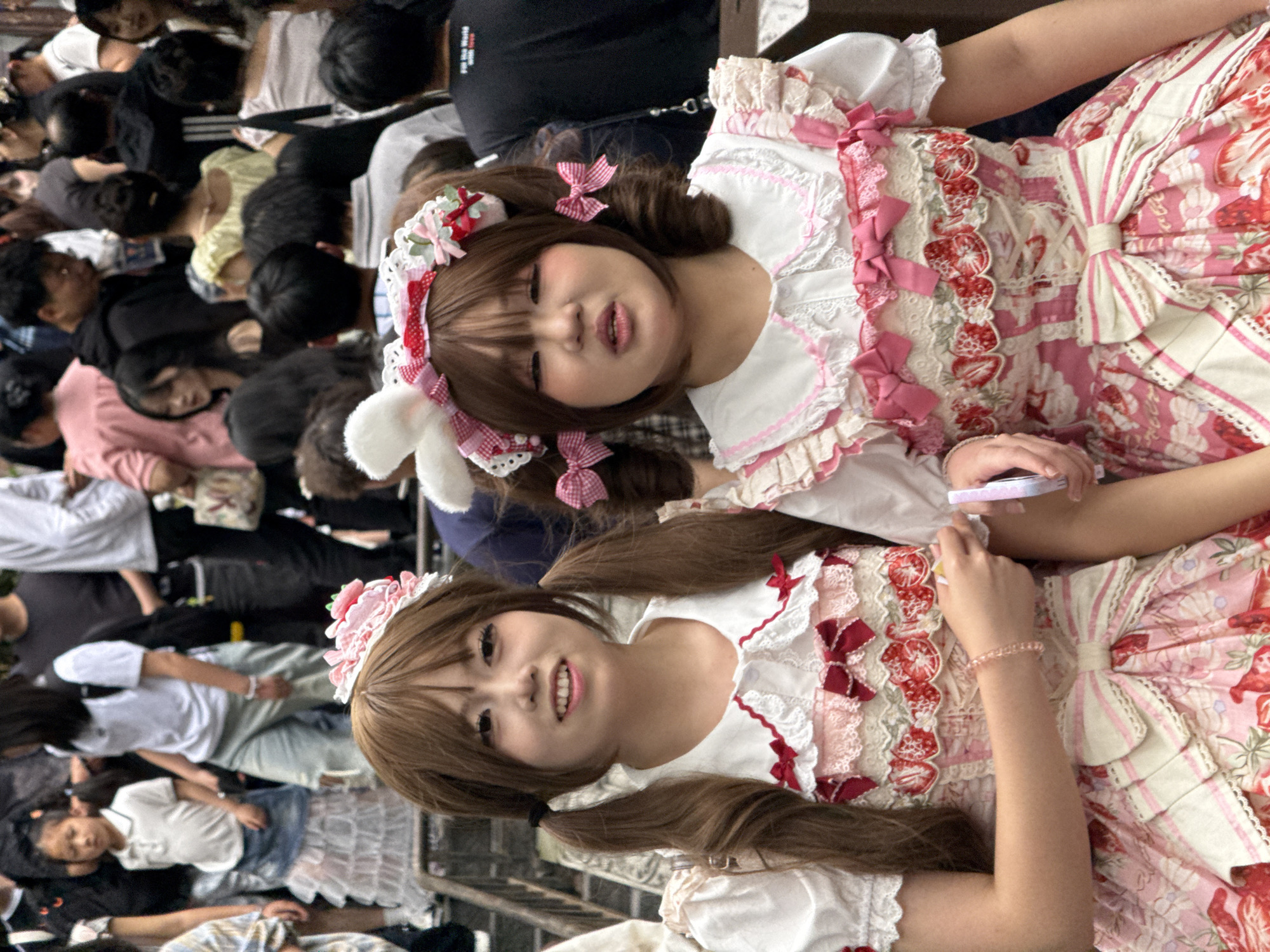 In Yuzhong District, China, two young women in elaborate Lolita-style dresses are the central subjects of the image. The dresses are pastel pink with white ruffles, bows, and strawberry patterns. One woman has brown hair styled in pigtails and wears a floral headband. The other has short brown hair with ribbons and a headband with bunny ears. A crowd of people surrounds them, some looking at the camera. The people in the crowd are dressed in casual clothing. The ground appears to be paved, and metal fencing is visible.
