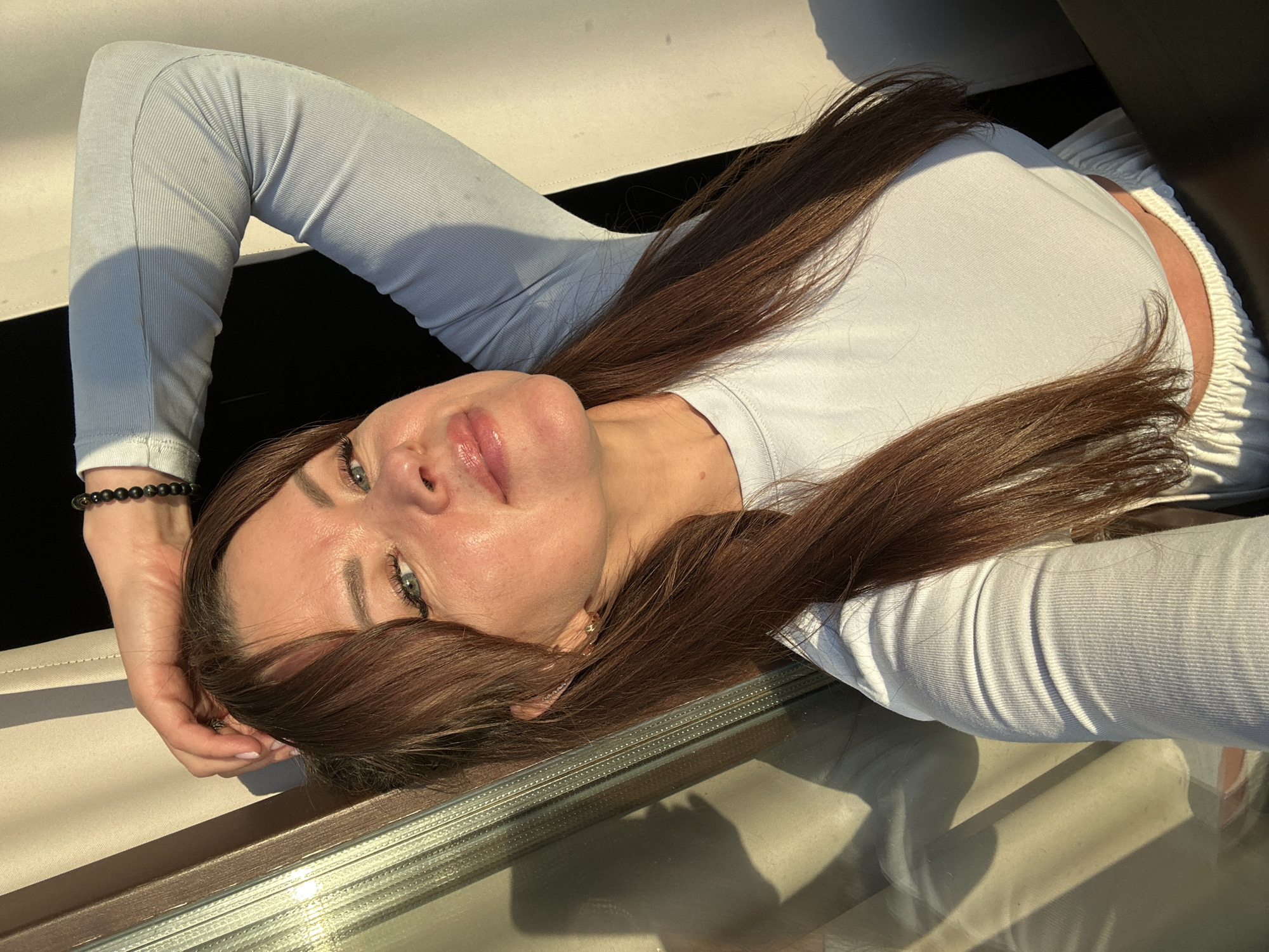 A woman is lying down, facing upwards and looking towards the camera. She has long, brown hair that is spread out around her head and shoulders. She is wearing a light blue, long-sleeved top and white pants. Her arms are bent, with one hand resting behind her head and the other near her face. She has blue eyes and her lips are slightly parted. There is a beaded bracelet on her left wrist. The scene appears to be outdoors, with sunlight casting shadows on the surface she is lying on, which seems to be a couch or lounge chair. There is a reflective surface in the foreground, possibly a table or window, showing the reflection of the scene and the woman. The lighting suggests it is daytime, with the sun creating a warm glow. The woman's expression is relaxed and serene.