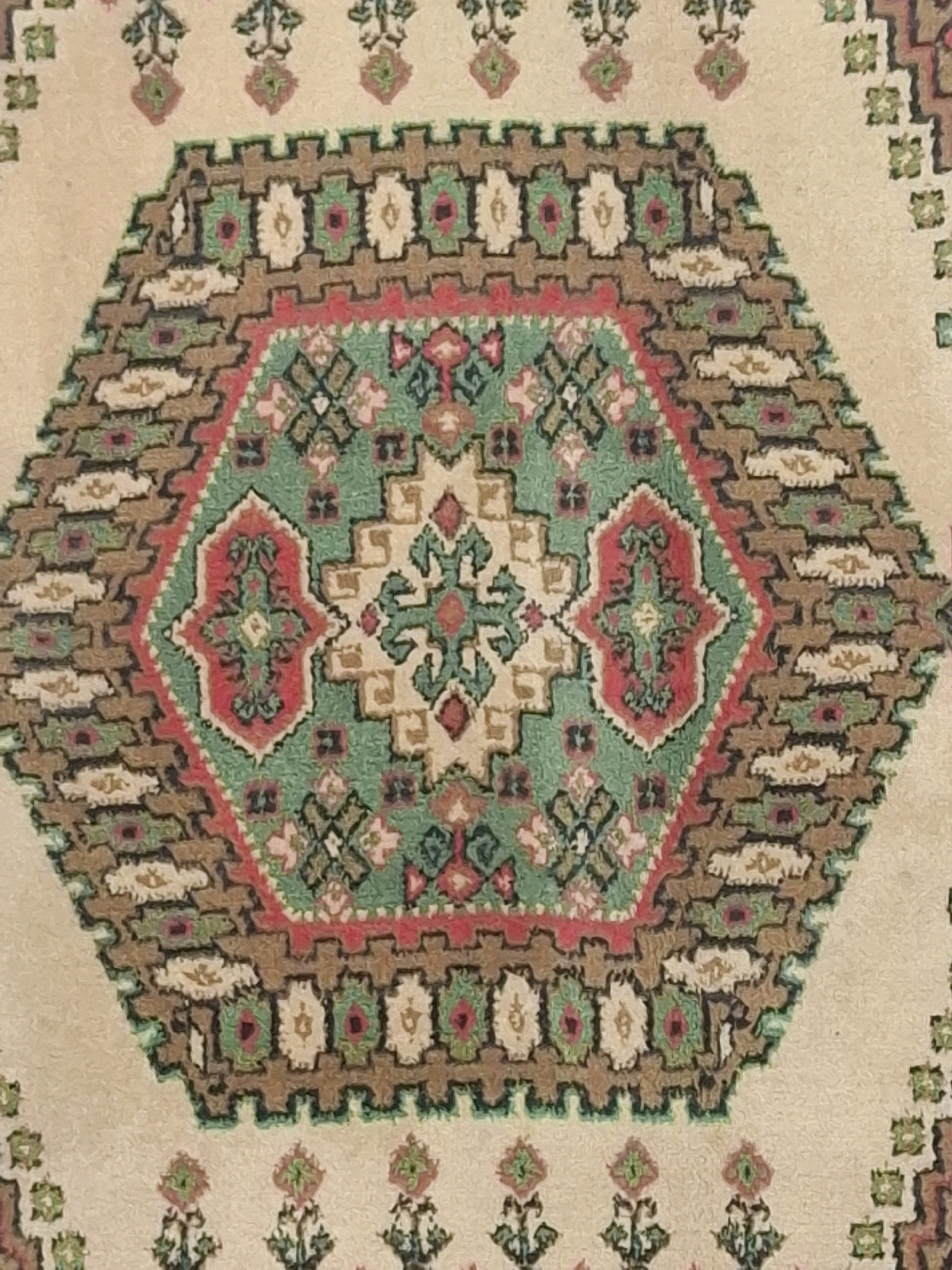 The image shows a close-up, top-down view of a Persian-style rug with a hexagonal central medallion. The rug features intricate geometric patterns in shades of beige, brown, green, and pink. The central medallion contains a stylized floral motif surrounded by smaller geometric shapes. The border of the medallion is a zigzag pattern in brown and pink, bordered by a green serrated edge. The background of the rug is a light beige color, with repeating small floral motifs scattered across it. There are no people or environmental elements visible in the image, it is a detailed view of the rug's pattern. The location of this rug in Almaty, Kazakhstan is not specified in the image itself and cannot be determined from the visual information provided.