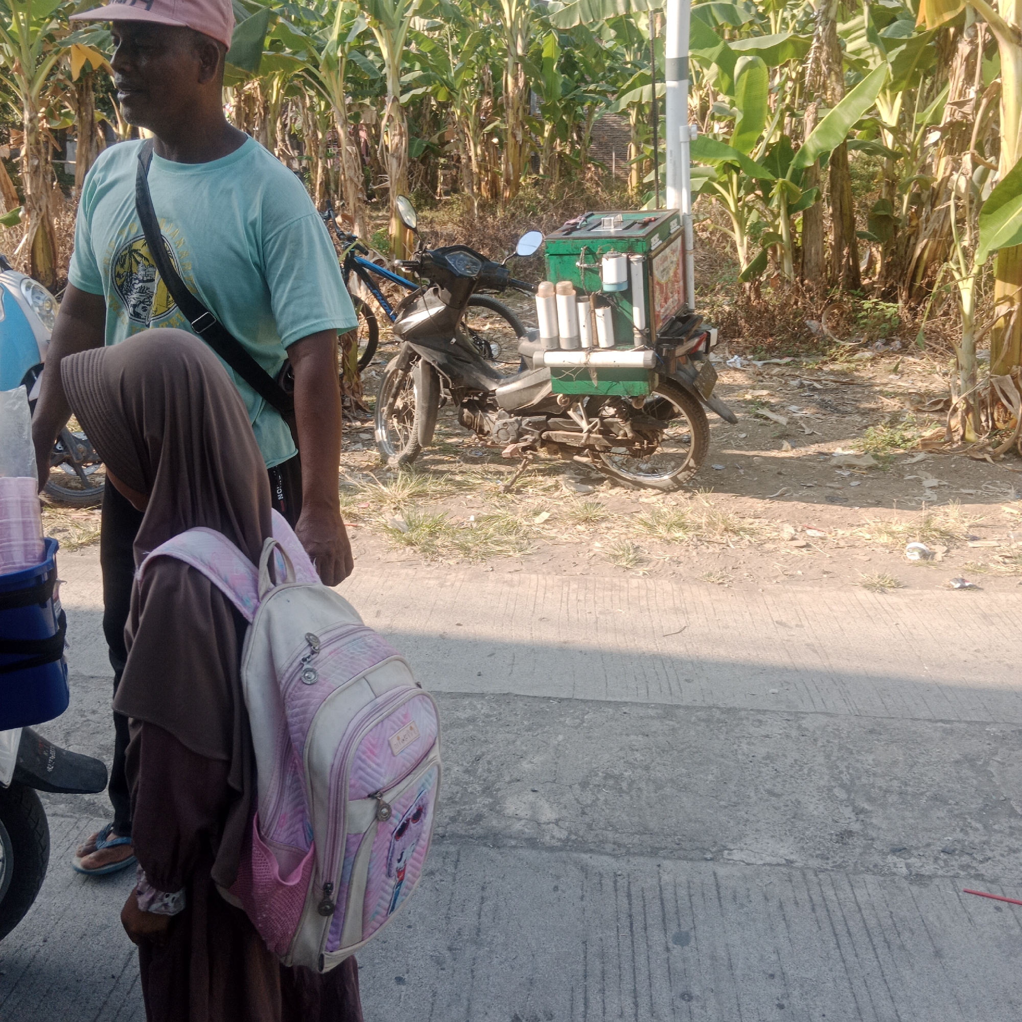The image depicts a scene on a street in Sidorejo, Indonesia, bordered by a dense grove of banana trees. A man wearing a light blue t-shirt and a pink cap stands on the left, with a dark strap across his chest. Beside him, to the right, a young person in a brown hijab with a pink and white backpack is visible. In the background, a motorcycle is parked, fitted with a green cart containing several white cylindrical containers. The ground is a mix of dirt and patchy grass, with a paved road surface in the foreground. The lighting suggests it is daytime, with sunlight casting shadows.