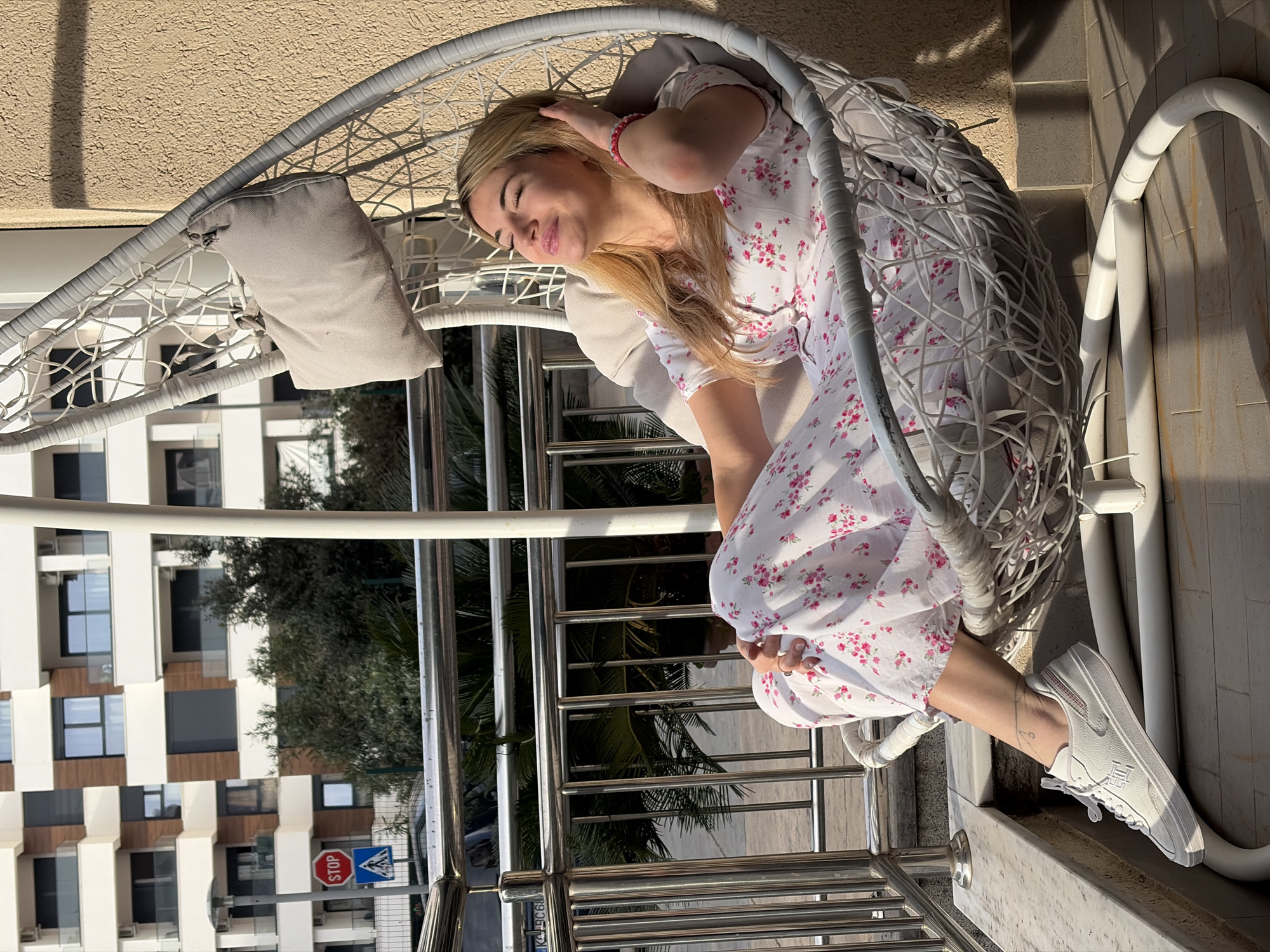 A woman with blonde hair wearing a white floral dress and white sneakers is sitting in a white wicker chair with a gray cushion. She is holding her hair with one hand and her eyes are closed, suggesting she is resting or sunbathing. The chair is positioned on a balcony with a metal railing. Outside the balcony, there is a view of buildings, trees, and a street sign. The buildings have a modern design with a mix of white and brown colors, and the street sign has a "STOP" sign and a pedestrian crossing symbol. The image is likely taken in Budva, Montenegro, based on the architectural style and general appearance of the surroundings.
