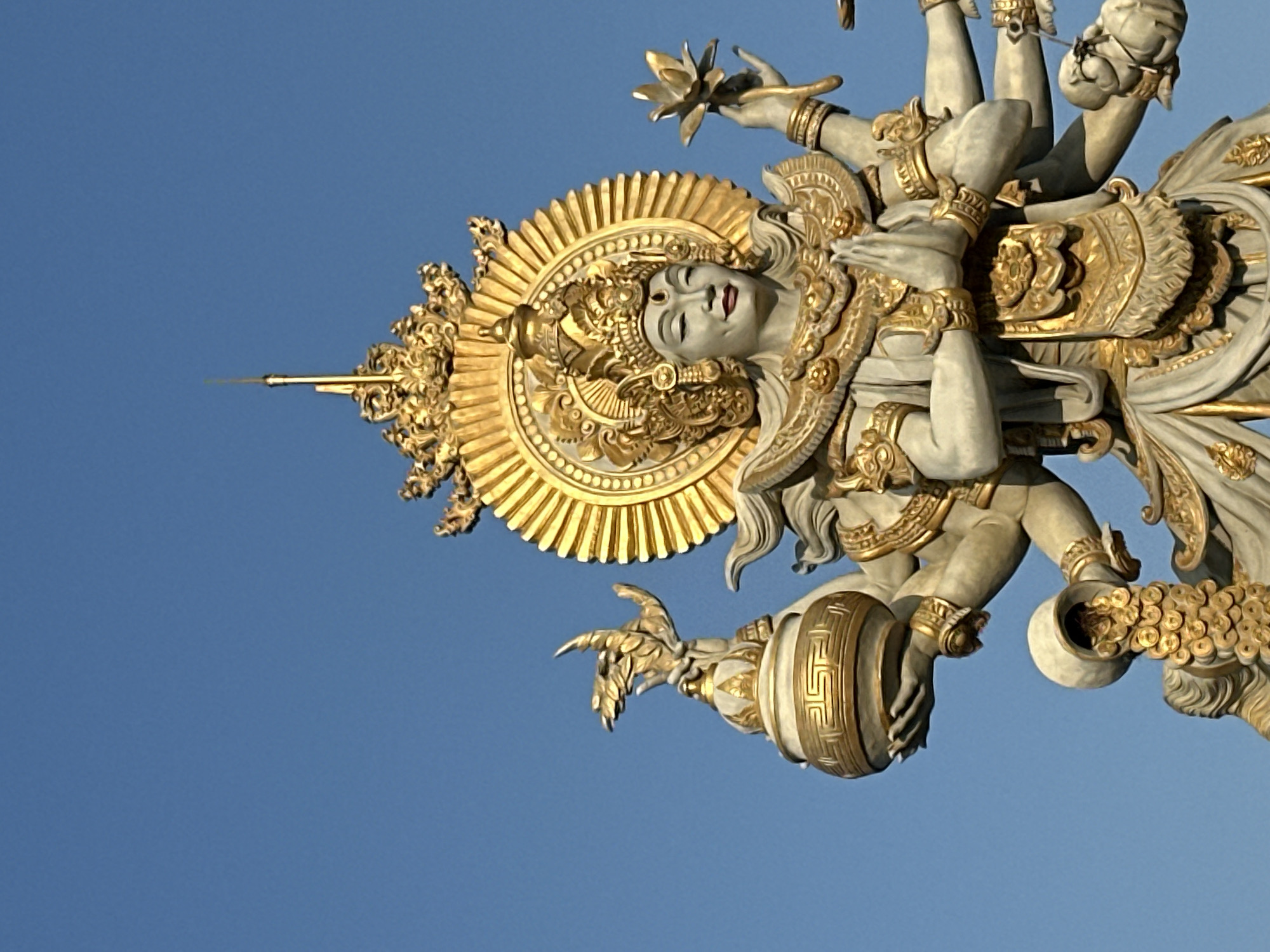 A large, ornate statue of a multi-armed deity stands against a clear blue sky. The statue is intricately detailed with gold accents and appears to be made of stone or concrete. The deity is depicted with its hands in a prayer position at its chest, and its other hands are holding various objects, including a lotus flower and a golden urn from which coins are pouring. The statue is adorned with elaborate headdresses and jewelry, characteristic of Indonesian religious art. The scene is likely located in North Kuta, Indonesia, a region known for its rich cultural heritage and numerous religious sites.