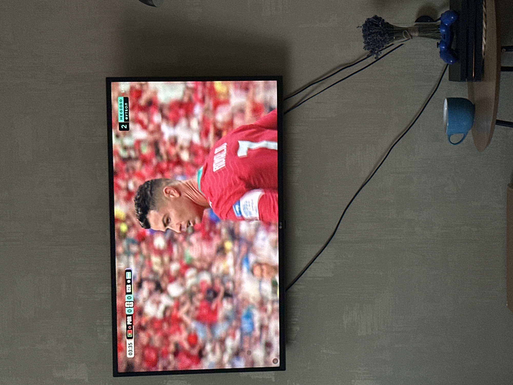 The image shows a television screen displaying a soccer match. The screen is oriented vertically, which is unusual for a TV. The match appears to be between Portugal (PUR) and an opponent, with the score currently 0-0. The time displayed is 03:35, and the word "LIVE" is visible in the top right corner of the screen, indicating the game is in progress.
The foreground of the screen shows a close-up of a soccer player wearing a red jersey with the number "1" on the back. The player's face is visible, and he seems to be looking down, possibly in concentration or disappointment. The background of the screen is blurred but shows a crowd of spectators, many of whom are also wearing red, suggesting they are supporters of one of the teams.
To the right of the television, a wall with a textured gray finish is visible. On this wall, a shelf holds a bunch of dried lavender and a blue decorative telephone. A blue mug sits on a wooden surface to the right of the shelf. Black cables are also visible running from the television towards the top right of the frame. The lighting in the room seems dim, and there are no strong indications of the weather or the time of day outside the context of the television broadcast. The overall scene suggests a casual setting, likely a home, where someone is watching a live soccer game.