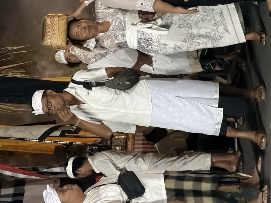 The image depicts a nighttime gathering of at least four adults participating in what appears to be a Balinese Hindu ceremony in Nusa Dua, Indonesia. In the center, an older adult male, wearing glasses, a white udeng (head covering), a white short-sleeved collared shirt with partially visible text "Dark Soul," a white patterned sarong (kamen), and brown sandals, is captured with his right hand near his mouth, possibly eating or gesturing. He carries an olive-green cross-body bag. To his immediate left, a partially visible adult male wears a white udeng, a white shirt, a plaid sarong, and flip-flops, carrying a black cross-body bag. To the right of the central male, an older adult female, with a visible mark on her forehead, wears a white lace kebaya, a white patterned sarong, and dark sandals. She carries a rectangular woven basket on her head, supported by both hands. Additional figures in traditional Balinese attire are partially visible in the background and foreground, including a male behind the central figure. The background features vertical banners or flags with red, white, and black patterns, along with ornate golden-hued structures, suggesting a ceremonial setting. The scene is illuminated by artificial light, casting shadows. The ground is a dark, flat surface.