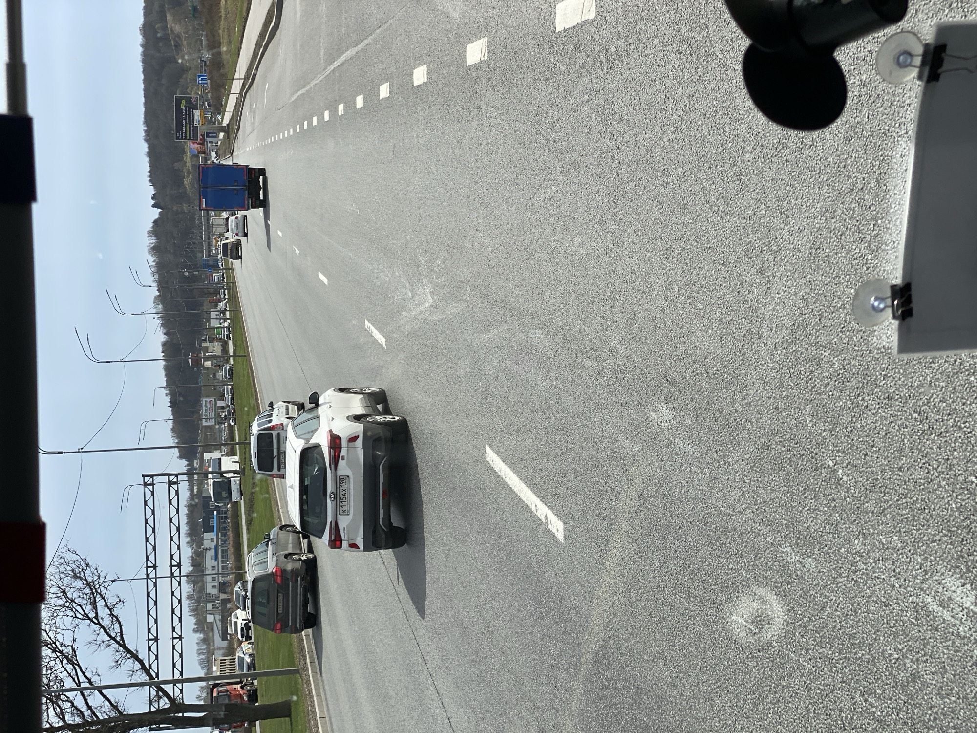 This media file captures a bright, sunny day scene on a multi-lane highway or major road in Saint Petersburg, Russia, viewed from inside a vehicle looking forward. The image itself is rotated 90 degrees clockwise.
On the asphalt road, which is marked with white dashed lane lines, several vehicles are present. A white Kia SUV, bearing the Russian license plate "K 115 AX 198" (with "198" being the region code for Saint Petersburg and Leningrad Oblast), is visible in the foreground, followed closely by a dark SUV. Further down the road, a blue truck with a box trailer is driving. Other cars and vans are parked or moving along the roadside and in the distance.
To the right of the road, a billboard with Russian text is clearly visible against a gentle hillside covered with sparse vegetation and bare trees. The text on the billboard reads "ТУРИСТИЧЕСКИЙ БУКЕТ 1.5-2" (Tourist Bouquet), with smaller, less legible text underneath. To the left, various infrastructure elements include bare trees, streetlights, and metal gantry structures, with distant buildings visible beyond them. The sky is clear and light blue.
In the immediate foreground, parts of the viewer's vehicle interior are visible on the right, including a dashboard or window frame, two suction cup mounts (one possibly holding a piece of paper), and a partial view of a dark, circular object, likely a camera or device. The bright sunlight casts distinct shadows from the vehicles, suggesting the middle of the day.
