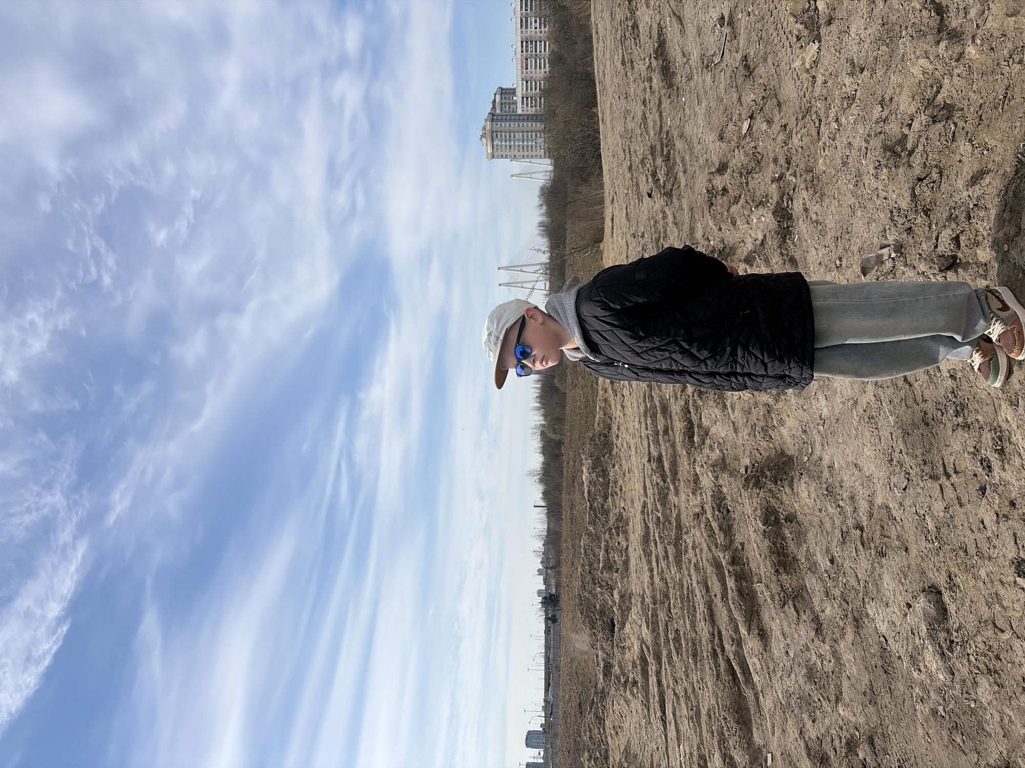A person wearing a baseball cap, sunglasses, black jacket, and light-colored pants stands on a dirt or sandy surface. In the background are buildings and a cable-stayed bridge, suggesting a location in Saint Petersburg, Russia. The sky is blue with streaks of white clouds.