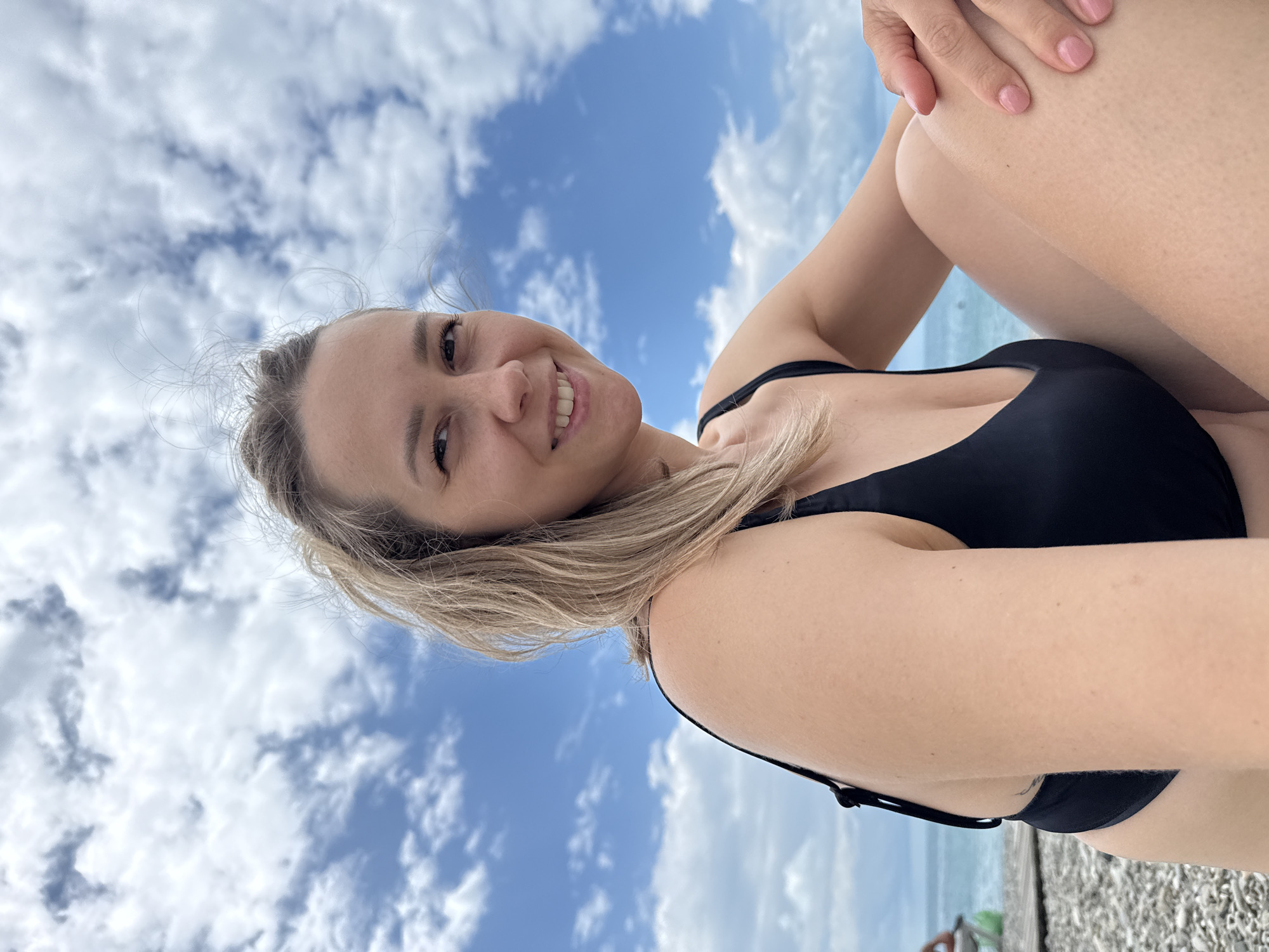 An outdoor, medium shot shows a fair-skinned Caucasian woman with blonde hair, wearing a black bikini, sitting on a pebble beach under a partly cloudy blue sky. The woman is looking towards the camera with a smile, her head turned slightly to the left. She has freckles on her face and her hair is loosely styled. Her left arm is resting on her lap, and her right hand is on her right leg. In the background, a body of water, likely the sea, is visible, with some clouds in the sky. The location is identified as Nebug, Russia.