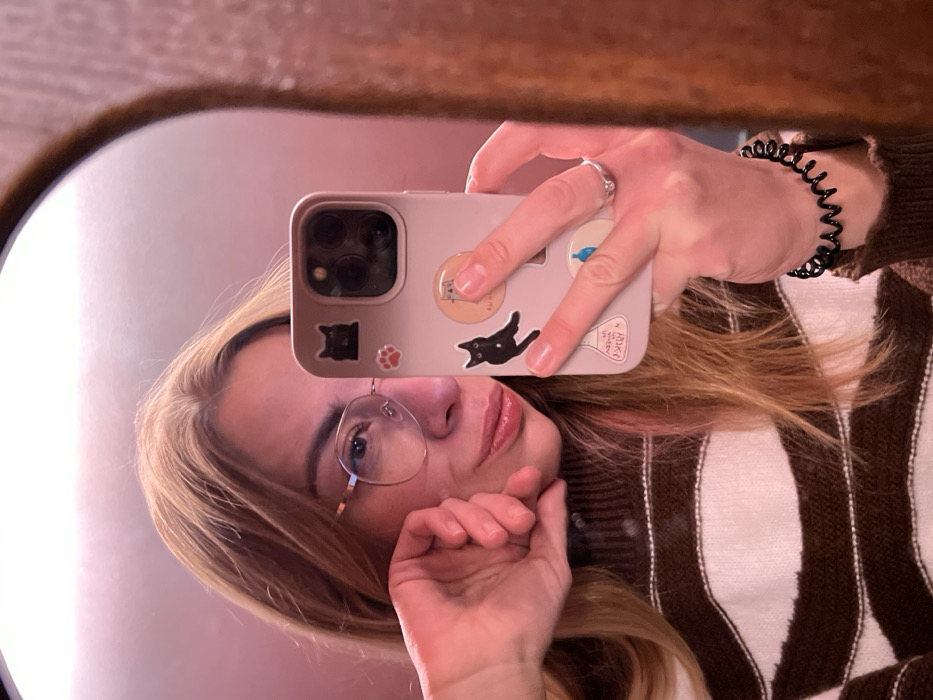 A female subject is depicted in a mirror selfie, captured within an indoor setting in Saint Petersburg, Russia. The subject has long, light-blonde hair and wears eyeglasses with thin frames. She is dressed in a brown and white horizontally striped high-neck sweater. Her left hand is positioned near her jawline, while her right hand holds a light pink-cased smartphone, identified as an iPhone Pro model by its camera array, pointed towards the mirror. The smartphone case is adorned with multiple stickers, including two black cats, a red paw print, a circular graphic, a beaker, and a blue liquid drop. A silver-toned ring is visible on her left ring finger, and a black coil hair tie is on her left wrist. The mirror features a dark, arched frame along the right edge of the image. The background reflected in the mirror is a uniform reddish-pink wall, suggesting warm, ambient lighting. The subject's lips are slightly pursed.