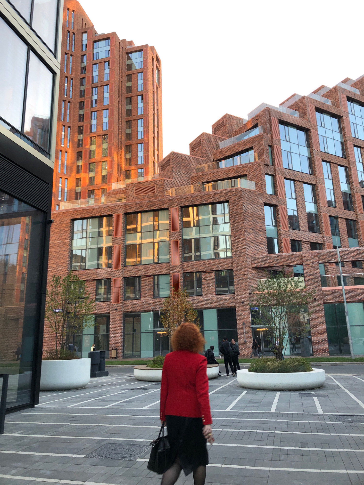 The image captures an outdoor urban scene, possibly in Moscow, Russia, featuring modern brick and glass architecture. In the foreground, a woman with curly red hair, wearing a red blazer and a black skirt, walks away from the camera, carrying a black handbag. Her back is to the viewer. The ground is paved with grey interlocking bricks, marked with white lines.
Dominating the background are several multi-story buildings constructed with red brick and large glass windows. The buildings exhibit a contemporary architectural style with stepped levels and visible balconies. Sunlight reflects off the glass facades, creating bright patches and distortions.
To the left, a dark-colored modern structure with reflective glass panels is partially visible. In the middle ground, there are large, white, curved planters with greenery. Several individuals are visible in the distance, interacting near the entrance of one of the buildings. Two men in dark clothing are standing and one is sitting on a bench, while others are walking or standing near the building's entrance. The overall impression is of a newly developed urban area with a mix of residential and commercial spaces. The sky is a pale, overcast white.