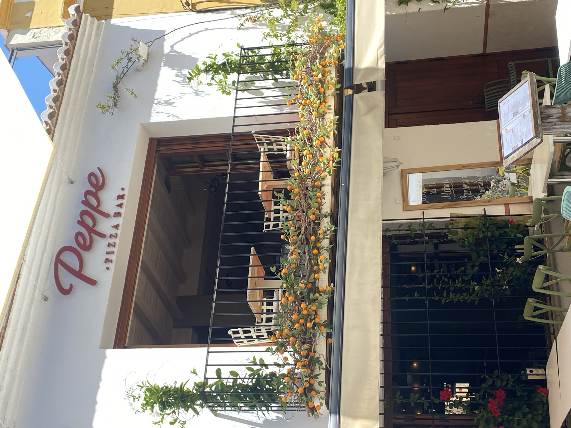The image shows the exterior of a restaurant in Nerja, Spain. The restaurant's name, "Peppe Pizza Bar," is displayed in red script on the white facade above a balcony. The balcony is decorated with artificial orange fruits and green foliage. Two tables and chairs are visible on the balcony. Below the balcony, a shaded seating area is visible, with tables and chairs, some of which are green. A menu stand is placed in front of a wooden door, and a tall mirror leans against the wall. The scene is bathed in sunlight, suggesting a daytime setting.