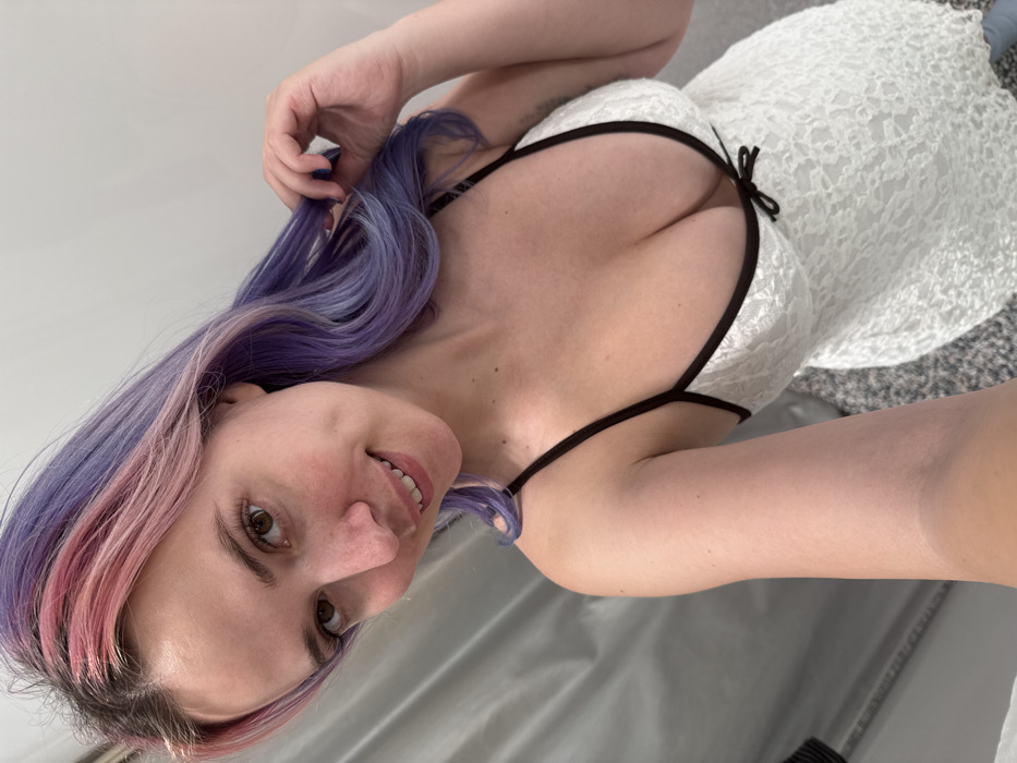 A female subject is depicted in a close-up, waist-up selfie within an unidentifiable indoor setting. The location cannot be precisely determined as being within the City of Zagreb, Croatia, based on the visual evidence, which suggests a private or commercial interior space such as a changing room. The subject has fair skin, brown eyes, and is looking directly at the camera with a subtle smile. Her hair is dyed, featuring a blend of pink and purple tones, with her natural darker roots visible at the scalp. She is wearing a white, textured or lace-patterned garment, possibly a camisole or short dress, with thin black spaghetti straps and a black trim along the neckline, which is deep and features a small black bow at the center. Her left hand is raised, with dark-painted fingernails (possibly blue), and her fingers are lightly gripping or styling a section of her purple hair near her head. The background consists of a plain, light-colored surface, possibly a wall or curtain, and a visible portion of a grey, textured floor, possibly carpeted. A dark, indistinct object is partially visible on the far left edge of the frame.