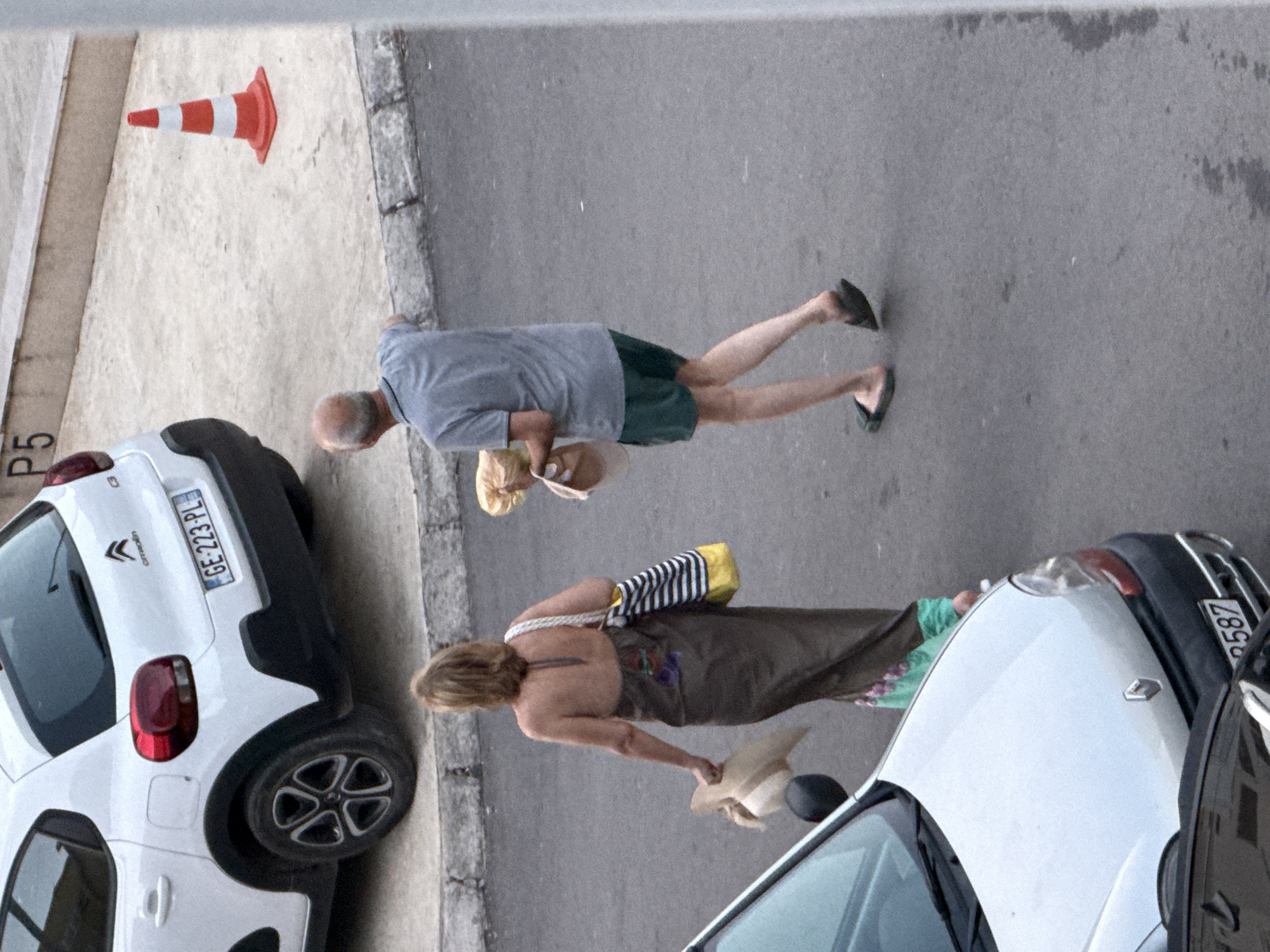 A top-down, medium shot captures a street scene in Budva, Montenegro. The ground is paved with asphalt and concrete. In the foreground, a white Renault car is partially visible, with its front hood and windshield showing. A black car is also partially visible in the bottom left.
Two individuals are walking away from the camera on the asphalt. The person on the left, a woman, is wearing a long, dark brown dress with a halter neck and appears to be carrying a beige hat in her left hand and a striped tote bag over her right shoulder. The person on the right, a man, is wearing a grey t-shirt and green shorts. He is carrying a brown paper bag in his left hand and what appears to be a white hat in his right hand.
In the background, a white Citroën car is parked. A red and white traffic cone stands to the right of the Citroën. A concrete barrier with the designation "P5" is visible behind the cone and the Citroën.