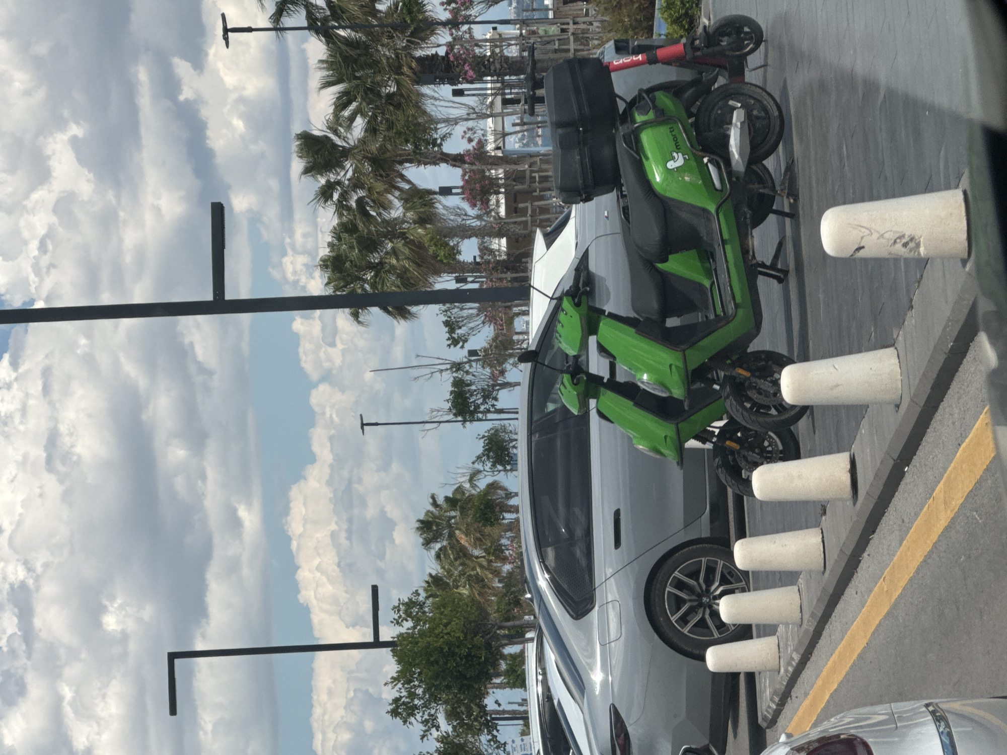 The image depicts a street scene in Konyaaltı, Turkey, characterized by a mix of urban and natural elements. The foreground features two parked electric scooters, one green and the other red. The green scooter is leaning against a silver car that is parked alongside a series of short, white concrete bollards demarcating a parking space along a yellow line on the asphalt.
Beyond the vehicles and bollards, palm trees are visible under a cloudy sky. Streetlights punctuate the scene. The overall environment suggests a coastal or near-coastal urban setting.