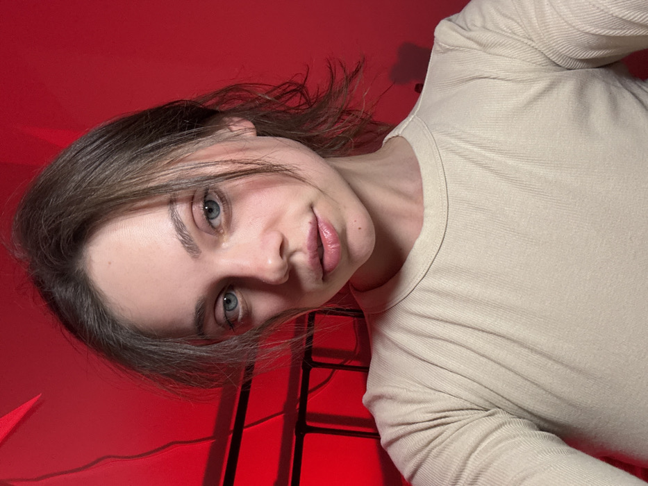 A young adult female is depicted in a close-up, head-and-shoulders shot. She possesses fair skin, light blue eyes, and medium-brown hair with strands framing her face. Her lips appear full with a subtle natural sheen. She is wearing a light beige or tan, ribbed long-sleeve crew-neck shirt. Her gaze is directed straight at the viewer with a neutral facial expression. The background is illuminated by a strong red light, creating an intense monochromatic environment. To the left, a dark, angular structure with horizontal bars is partially visible, suggesting a piece of furniture such as a bed frame or shelving unit. A lighter, sharp-edged element is present in the upper left corner. The setting is an indoor environment. The specific location within the city of Fiuggi, Italy, cannot be determined from the visual information provided in the image.