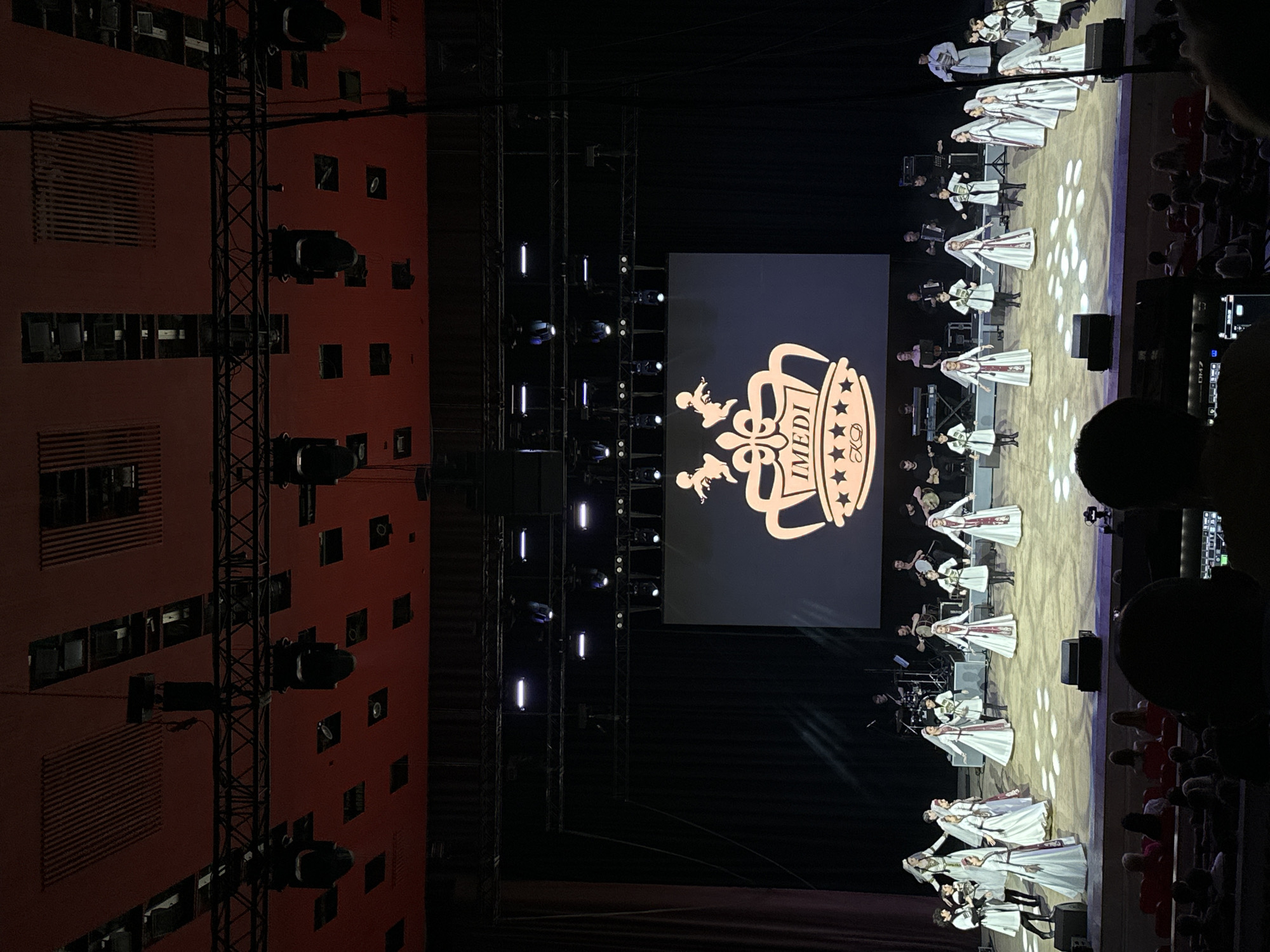 The image depicts a performance on a stage, likely in Sochi, Russia, given the provided context. A group of performers, predominantly young women and girls dressed in traditional white and red attire with headscarves, are arranged on the stage. They appear to be in motion, possibly a dance or choreographed performance. Behind them, a band is visible, with individuals playing instruments such as accordions and drums, contributing to the live music. A large screen at the back of the stage displays a logo that includes a crown and the word "IMEDI," suggesting a cultural event or a branded performance. The stage is illuminated by spotlights, casting patterns on the floor. In the foreground, the audience is partially visible, seated in a dimly lit auditorium, with some audience members holding phones, likely recording the event. The overall environment suggests a formal performance setting within an entertainment venue.