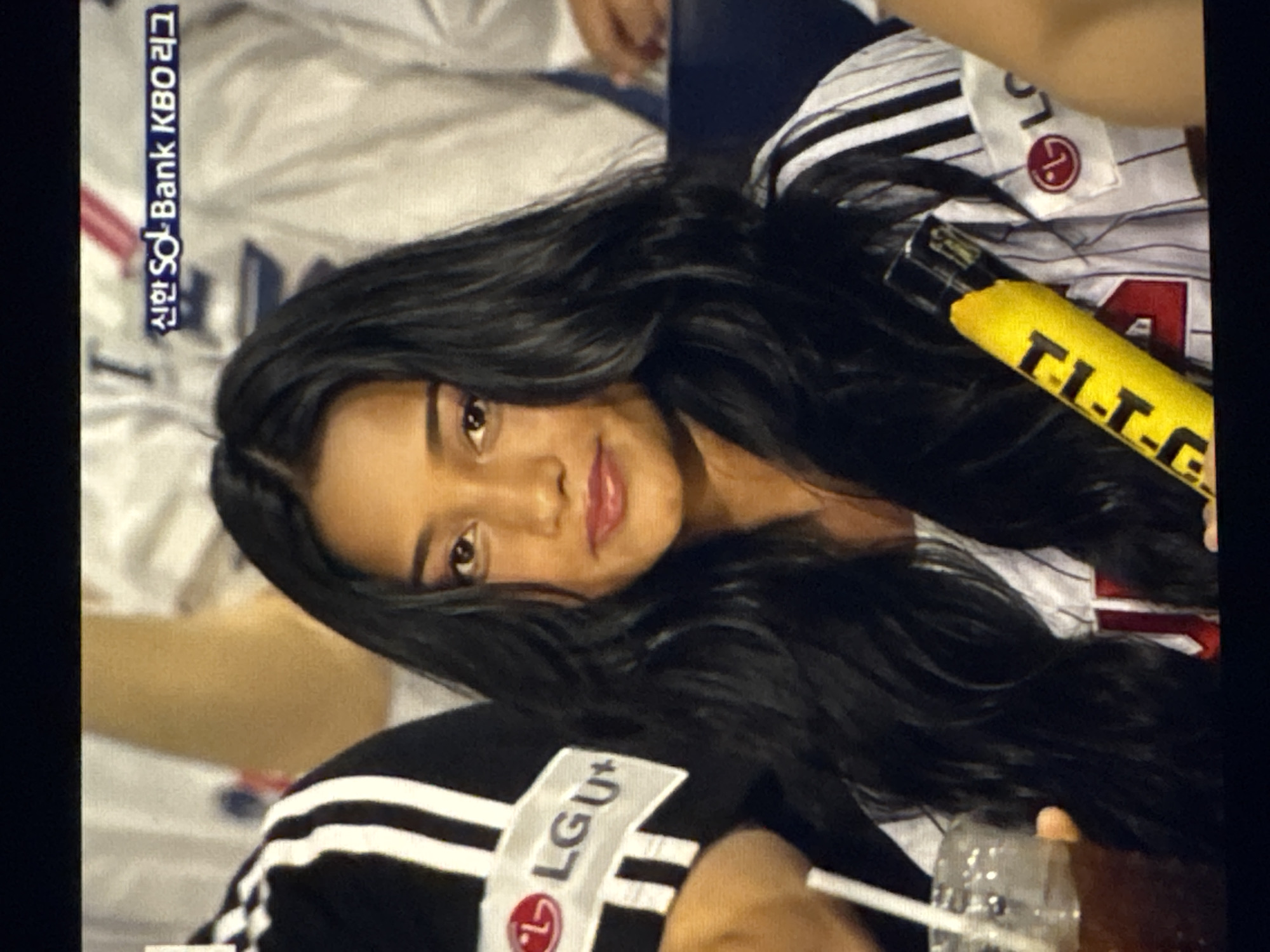 The image shows a close-up of a young woman with long, dark, wavy hair. She is wearing a white and black striped jersey with red and white lettering, and a patch with the LG logo. She is holding a yellow foam cheering stick in her right hand. Her left hand is visible holding a clear plastic cup with a straw containing a dark liquid. In the background, there is blurry text that appears to be in Korean, and other people in sports attire. There is no indication that this image was taken in Kaunas, Lithuania.