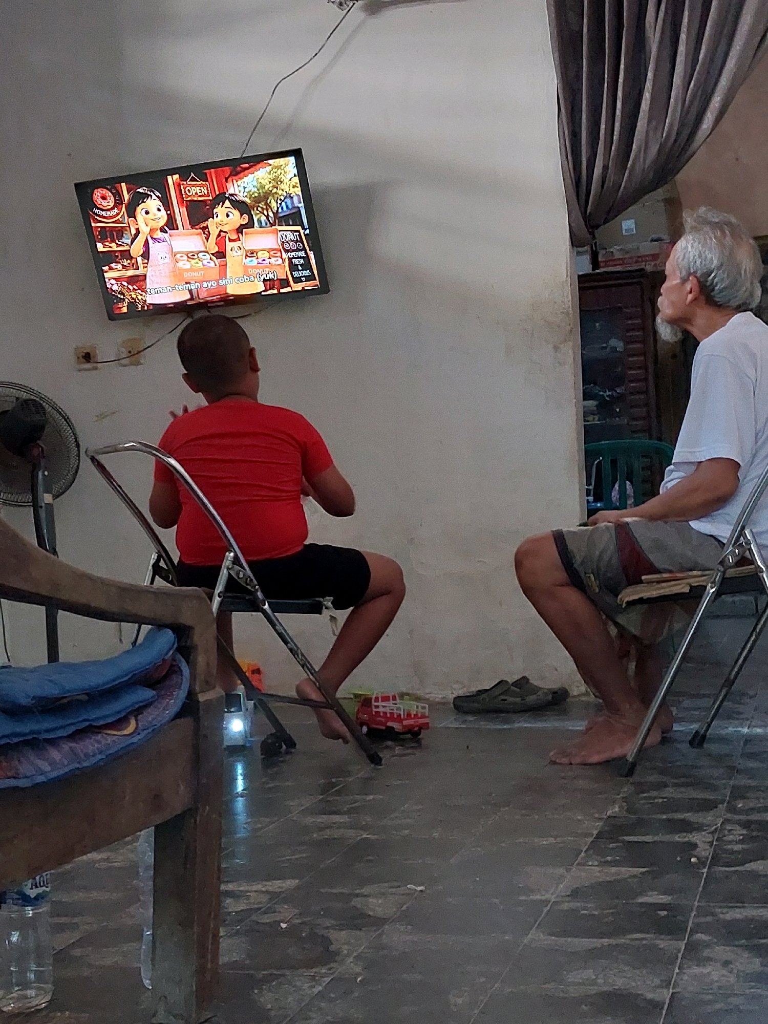 In SARANGAN, Indonesia, an indoor scene depicts a television screen displaying an animated show with two young girls, one wearing an apron and holding donuts. In front of the television, a young boy with a shaved head, wearing a red t-shirt and black shorts, is seated on a metal folding chair, facing away from the camera and gesturing with his hands. To the right, an elderly man with white hair and glasses, wearing a white t-shirt and patterned shorts, sits on another metal folding chair, facing the television and looking upwards. The room has a concrete floor, a partially visible fan on the left, and a dark curtain hanging on the right. A stack of blue cushions is visible on a wooden structure on the left. Several toy vehicles are on the floor.