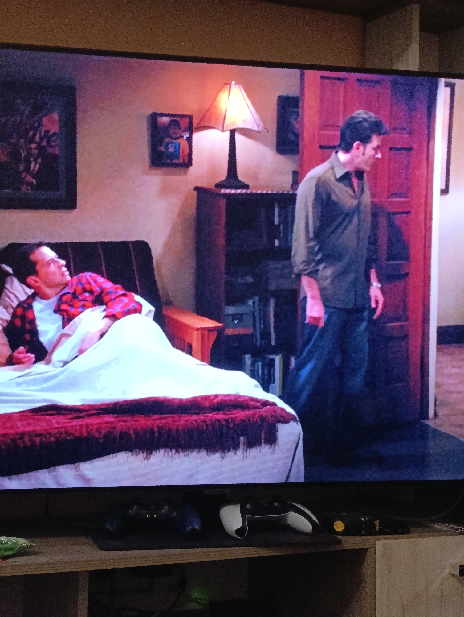 The image shows a television screen displaying a scene from a TV show. A man in a red and blue plaid shirt lies in a bed with a white sheet and a dark red blanket. Another man stands near a partially open wooden door. Behind the bed is a dark wooden bookcase with books and objects. A lamp sits on top of the bookcase. On the wall, there are three framed images. The image does not show any information that indicates the location is in Plovdiv, Bulgaria. The scene appears to be set indoors, likely in a bedroom. Below the television are game controllers and other electronic devices.