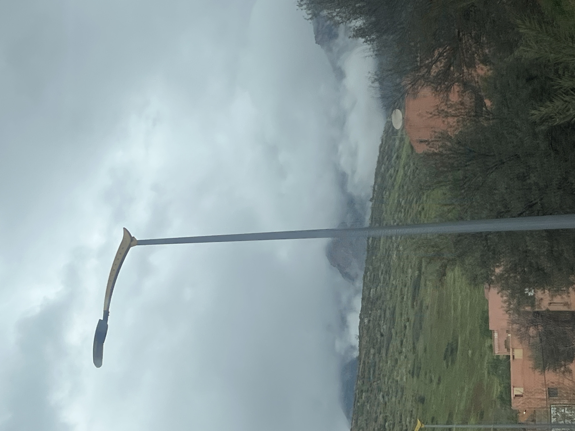 The image captures a scene in Ouaouizert, Morocco, under an overcast sky filled with dense, grey clouds that partially obscure the rugged mountain peaks in the background. A tall, grey metallic streetlight pole with a yellow-orange, curved arm and a dark luminaire extends vertically through the center-left of the frame. To the far left, a portion of another similar yellow streetlight arm is visible. A verdant, green hillside with undulating terrain rises behind the streetlights. In the midground, several light reddish-brown, multi-story buildings are partially visible through dense, dark green shrubbery and trees. One building on the right features a white satellite dish mounted on its roof. No individuals or specific actions are discernible within the frame.