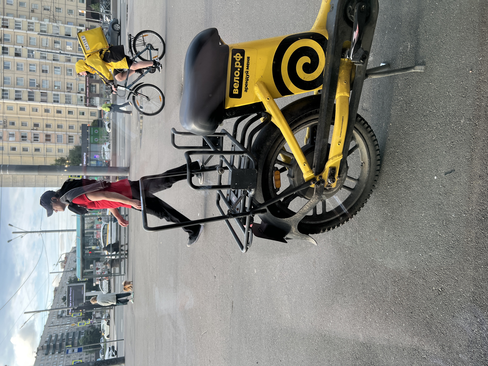 In the city of Sinyavino, Russia, a person on a bicycle with a yellow delivery bag navigates a paved road. The person is wearing a yellow hooded jacket. In the foreground, a yellow electric bicycle is parked on a kickstand. The bicycle has a black seat and a black cargo rack. Text on the bicycle reads "вело.рф" and "арендуй меня". In the background, a person walks across the street, carrying a sweater over their shoulders. Other pedestrians and buildings are visible in the distance. The sky is cloudy.