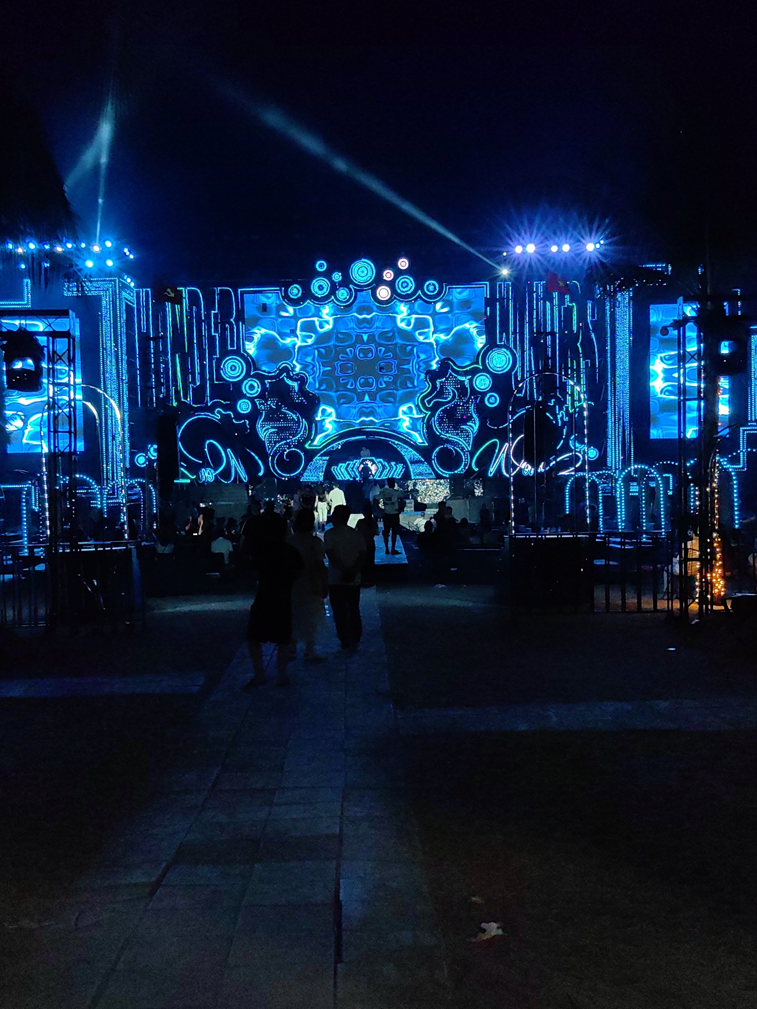 The image shows an outdoor event at night in Bai Chay Ward, Vietnam. A large stage is illuminated with blue lights, featuring geometric patterns and designs. Several people are visible in the foreground and middle ground, some walking towards the stage. Tables and chairs suggest a seated viewing area. A stone pathway leads towards the stage. The scene suggests a concert, festival, or similar large-scale entertainment event.