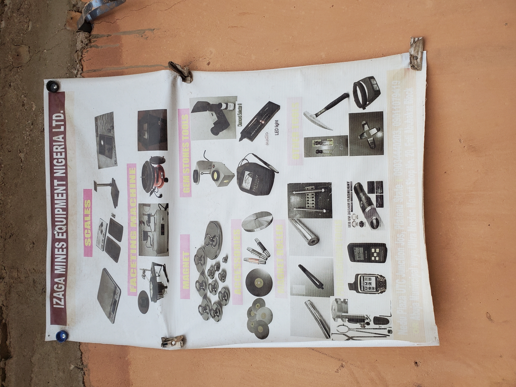 A large, white poster with the title "IZAGA MINES EQUIPMENT NIGERIA LTD." is affixed to a textured, orange-brown wall. The poster displays various mining and gemological equipment, categorized under headings such as "SCALES," "FACETING MACHINE," "MAGNET," "GEM STONES TOOLS," "ACCESSORIES," and "OFFICE TOOLS." Each category features photographic illustrations of different items. At the bottom of the poster, contact information is visible, including a phone number (09034440203, 09011079419) and an address: "Nedu Plaza UTC Junction Jos, Plateau State" and "Shop No. 20, Fati Abubakar Block, Abuja Municipal waye Ultra Modern Market." The poster is secured to the wall with blue and dark-colored circular fasteners, and a metal clip is visible on the left side. The environment appears to be outdoors, with rough textures on the wall and parts of the poster showing wear and tear, suggesting it has been exposed to the elements.