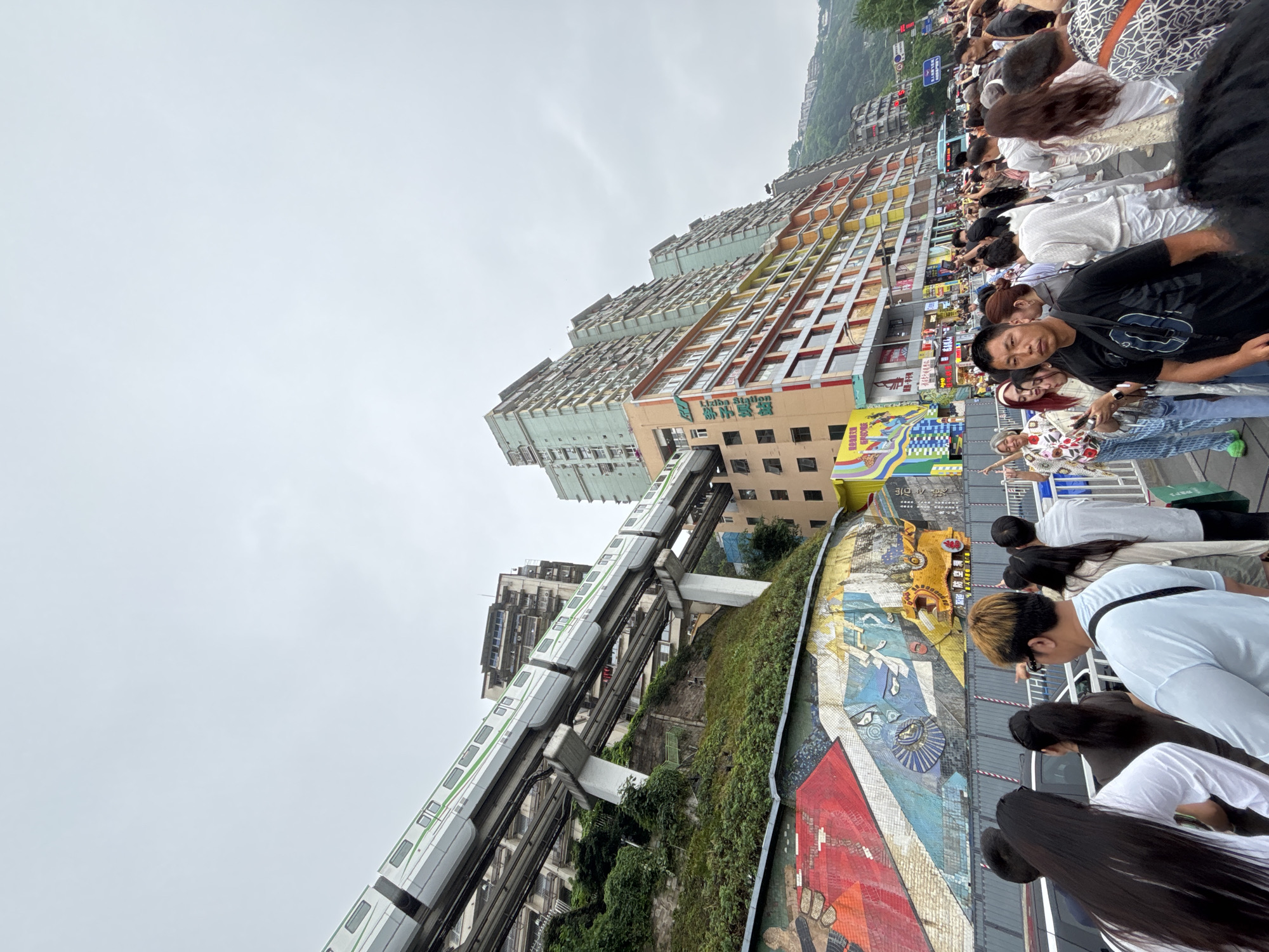 The image shows a crowd of people looking at a monorail train as it passes by a building in Yuzhong District, China. The building is tall and has many windows, and there is a large mural on the side of the building. The sky is overcast, and the surrounding area is urban.