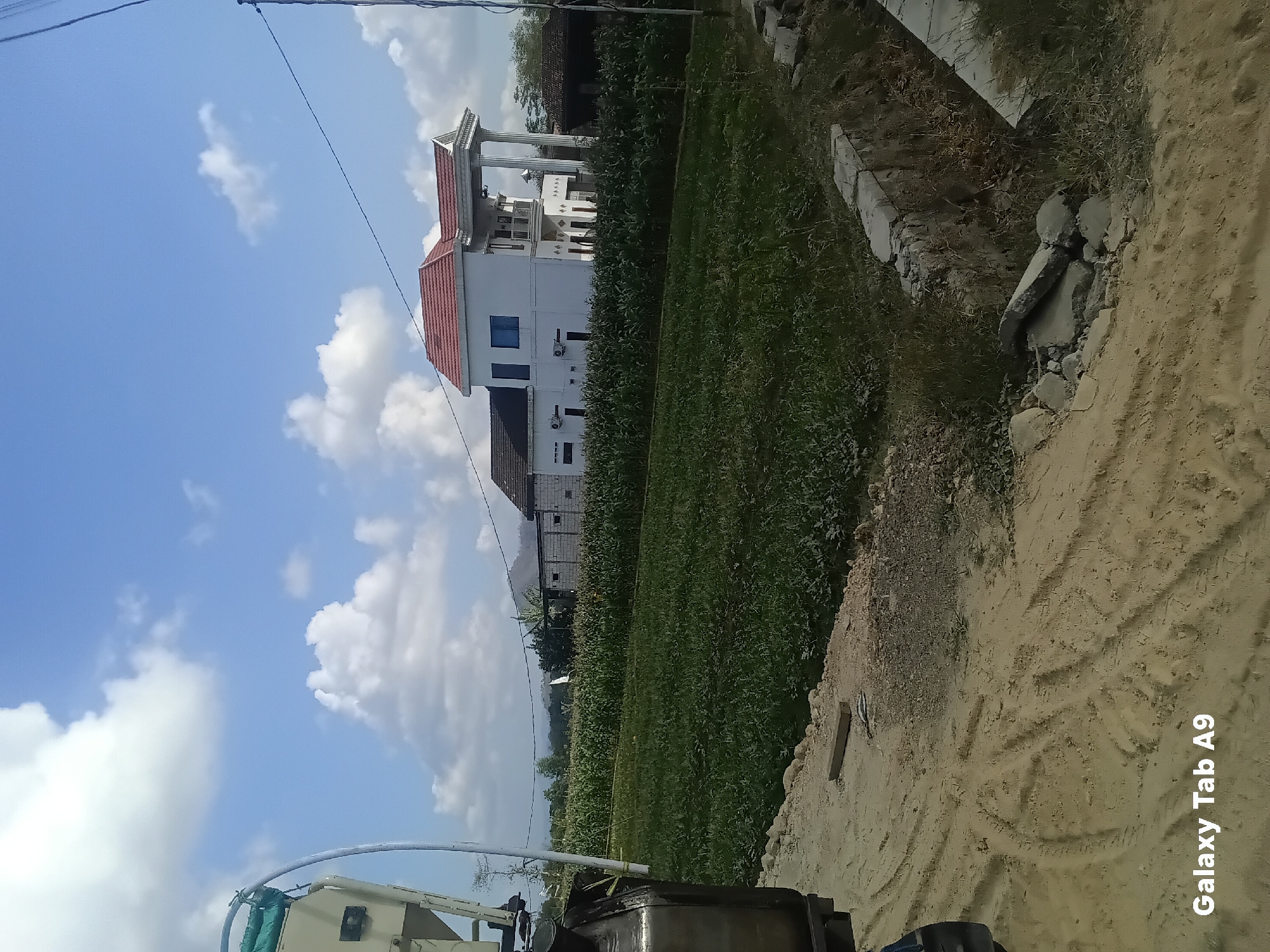 The image depicts a rural scene in Bendorejo, Indonesia, under a bright, partly cloudy sky. In the foreground, a dirt path shows tire tracks and scattered debris. To the left, part of a large, metallic agricultural machine is visible. Beyond the path, a field of tall green crops, likely corn, stretches towards the middle ground. In the middle ground, a modern, multi-story white house with a red-tiled roof stands prominently, featuring large columns and balconies. Adjacent to it, other buildings with brick and tiled roofs are present. The background consists of trees, distant hills, and the sky. The overall impression is one of a developing rural area with both agricultural activity and modern residential structures.