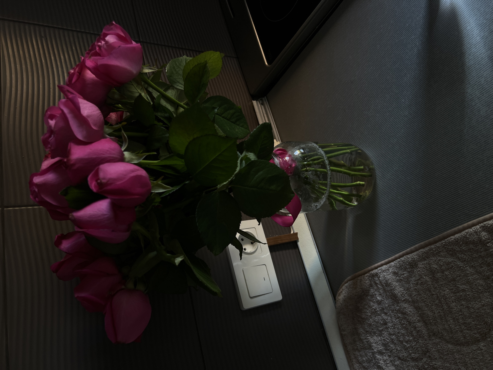 In a domestic kitchen setting in Tarasovka, Russia, a bouquet of bright pink roses is arranged in a clear glass vase filled with water. The vase rests on a gray countertop, likely part of a kitchen island or counter. To the left of the vase, a beige textured mat or towel is partially visible. Behind the roses, a white electrical outlet with a switch is mounted on a dark, textured wall. The scene is illuminated by natural light casting shadows and highlighting the glossy petals of the roses and the smooth surface of the countertop. A portion of a black stovetop is visible on the right side of the image.
