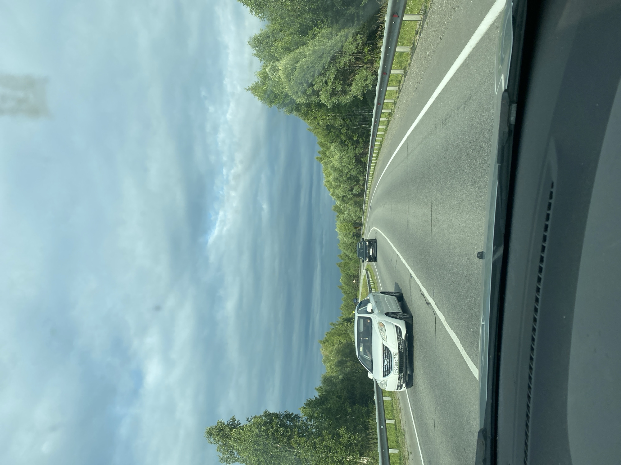 The image shows a view from inside a vehicle, looking forward and to the side. The perspective is slightly tilted. A paved road with a white line marking the lane curves to the right. A metal guardrail runs alongside the road, separating it from a grassy shoulder. Dense green trees line the other side of the road, forming a natural barrier.
On the road, there are at least two vehicles visible. A white SUV with a license plate that reads "C95ВЕС 775" is in the foreground, to the right of the lane. Further ahead, a dark-colored car is also on the road.
The sky above is filled with dramatic clouds. Some parts are bright and blue, while others are dark and ominous, suggesting an approaching storm or the aftermath of one. The lighting indicates it is daytime, likely late afternoon or early evening, given the contrast between the bright and dark clouds.
The overall scene is one of travel along a highway bordered by natural greenery, under a dynamic sky. The location is specified as Aisha, Russia. There are no people clearly visible within the vehicles or outside. The emotional tone of the image is neutral, focusing on the landscape and the act of driving.