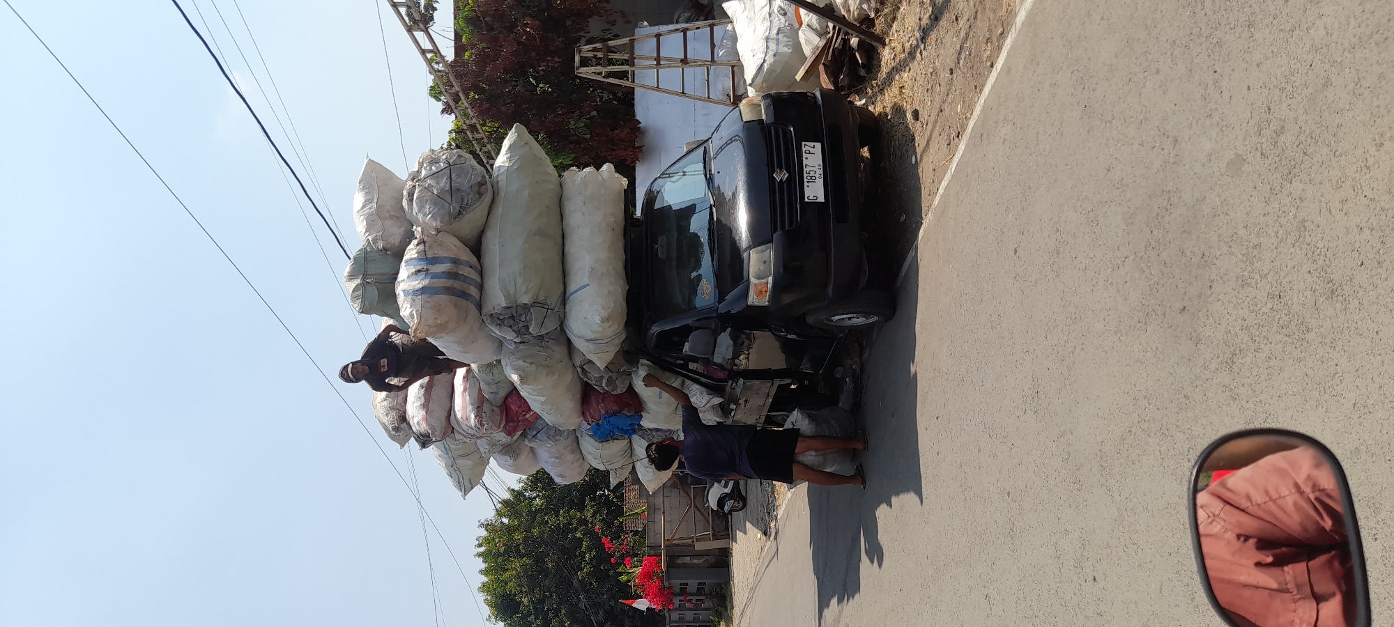 In the city of Mengori, Indonesia, a black Suzuki carry pickup truck is heavily overloaded with large sacks of goods. Two men are visible; one, wearing a dark hooded shirt and dark pants, stands on top of the sacks, appearing to be securing them. Another man, wearing a dark t-shirt and dark shorts, stands beside the truck, reaching up towards the sacks. The sacks are stacked precariously high, exceeding the capacity of the truck. The scene is set on a paved road, with a sidewalk and sparse vegetation visible on the right side of the road. Power lines stretch across the clear blue sky overhead. A motorcycle is parked in the background, and a ladder is leaning against a white structure. The image appears to be taken from a low angle, possibly from another vehicle.