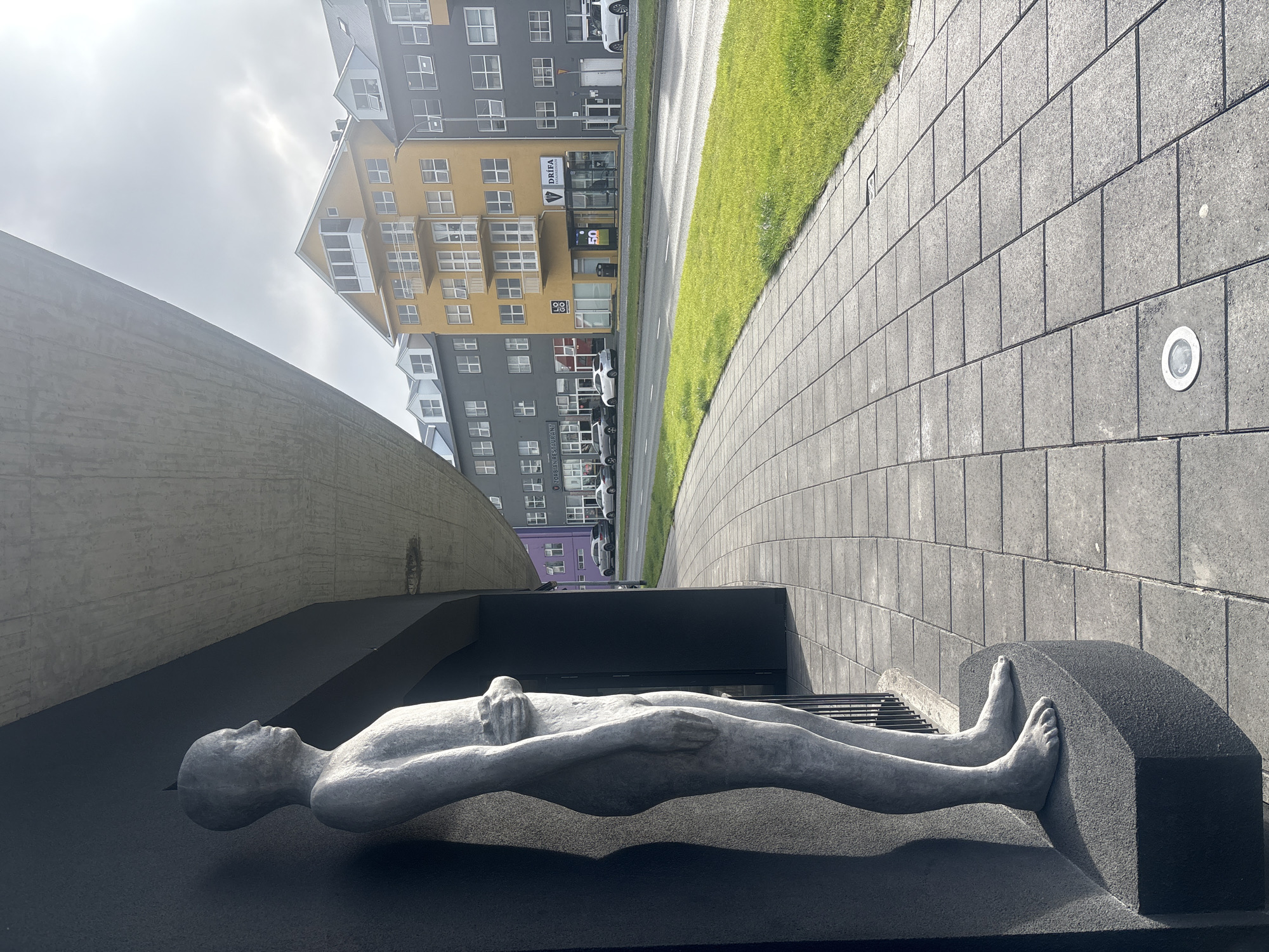 The image depicts a sculpture of a male figure standing on a dark grey pedestal, positioned to the left of a grey tiled sidewalk. The sculpture is grey and textured, with the figure's arms crossed over its chest. Behind the sculpture, a dark, textured wall forms the exterior of a building. The sidewalk extends to the right, leading towards a street lined with colorful buildings. The buildings are of varying heights and colors, including yellow, grey, and purple. The yellow building in the center has multiple floors with balconies and numerous windows. Across the street, there is a grassy median strip and a road with several cars. The sky is overcast with patches of sunlight. This scene is located in Reykjavik, Iceland, specifically on a street with commercial buildings and pedestrian walkways.