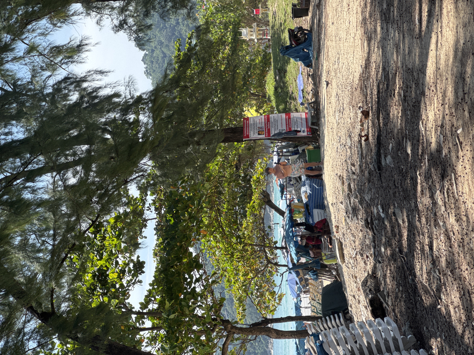 The image shows a beach scene in Karon, Thailand. In the foreground, there is sandy ground with scattered debris and shadows from trees. In the midground, a man is standing and looking at his phone. Behind him, there are several beach umbrellas, lounge chairs, and bags, suggesting a vendors' area. A sign with text in Thai and English is posted on a tree. To the right, a motorcycle is covered with a blue tarp, and a small shrine-like structure is visible in the background. Beyond the beach setup, the ocean with waves can be seen, and lush green hills form the backdrop. The sky is clear and blue.