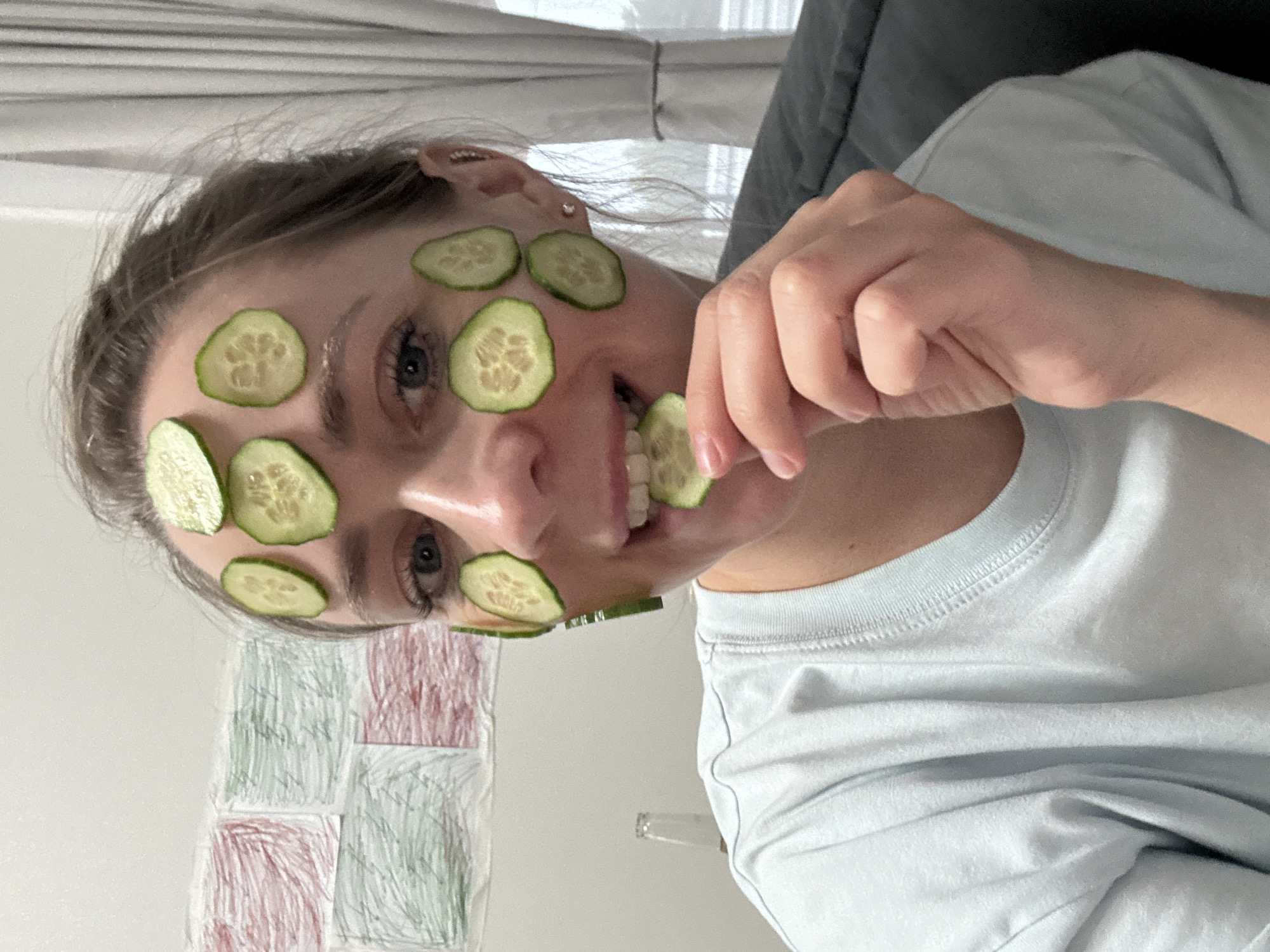 A young woman is lying on her back, wearing a light blue t-shirt and sporting cucumber slices on her forehead, eyes, cheek, and mouth. She holds a cucumber slice up to her mouth with her hand. A grey couch is behind her head. A piece of paper with green and red crayon squares is beneath her face. The photo was taken indoors with natural light. The location is not discernable as Chaikovsky, Russia.