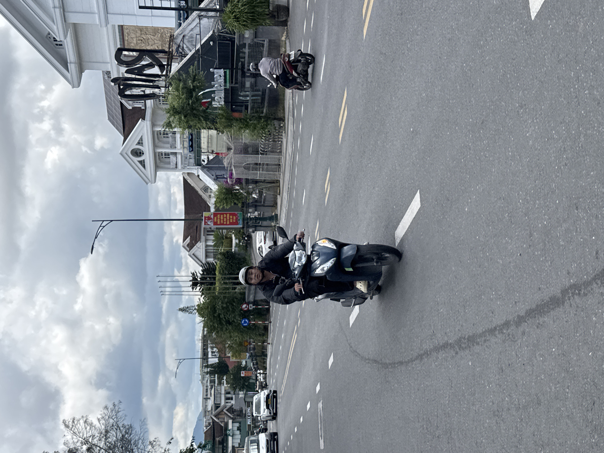 The image captures a street scene in Da Lat, Vietnam, likely on a cloudy day. In the foreground, a person wearing a dark jacket, light-colored pants, and a white helmet rides a dark-colored scooter. The scooter has a camera attached to its handlebars. The rider appears to be looking forward. The street is paved with asphalt and marked with white dashed lines. There is a slight curve in the road.
In the background, several buildings with white facades and sloped roofs are visible. One building has a prominent sign with large, dark letters that appear to spell "RA." Another building has a Heineken logo. Trees and vegetation are interspersed between the buildings. Further down the street, other vehicles, including a white van and a white car, are present. The sky is overcast with visible clouds. The overall impression is a typical urban street in a Vietnamese city, with a focus on the activity of the scooter rider.