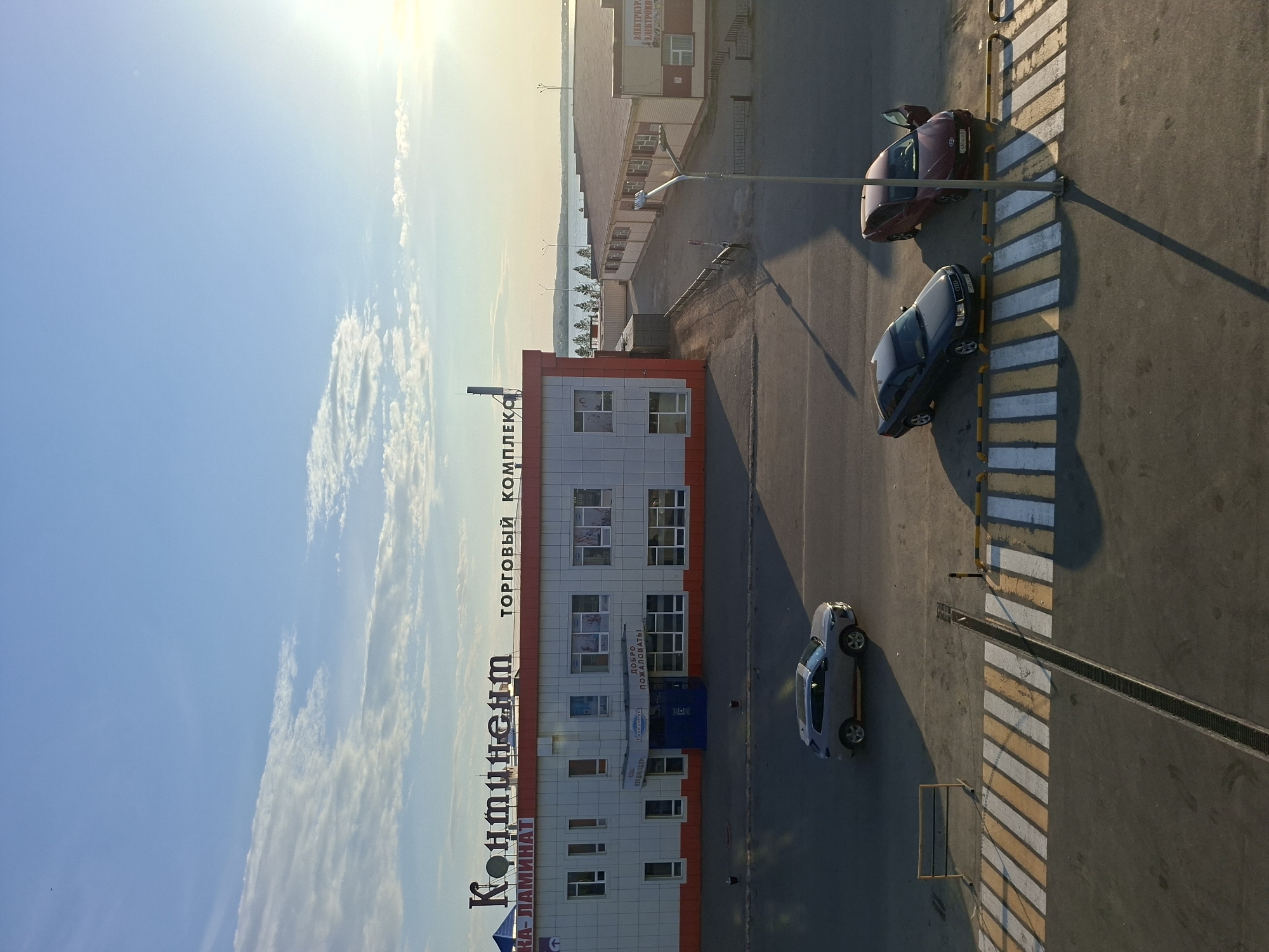 The image shows an aerial view of a street scene in Kokshetau, Kazakhstan. A building labeled "КОНТИНЕНТ" and "ТОРГОВЫЙ КОМПЛЕКС" is present. Three cars are visible on the street near a pedestrian crossing. The sky is blue with some clouds.