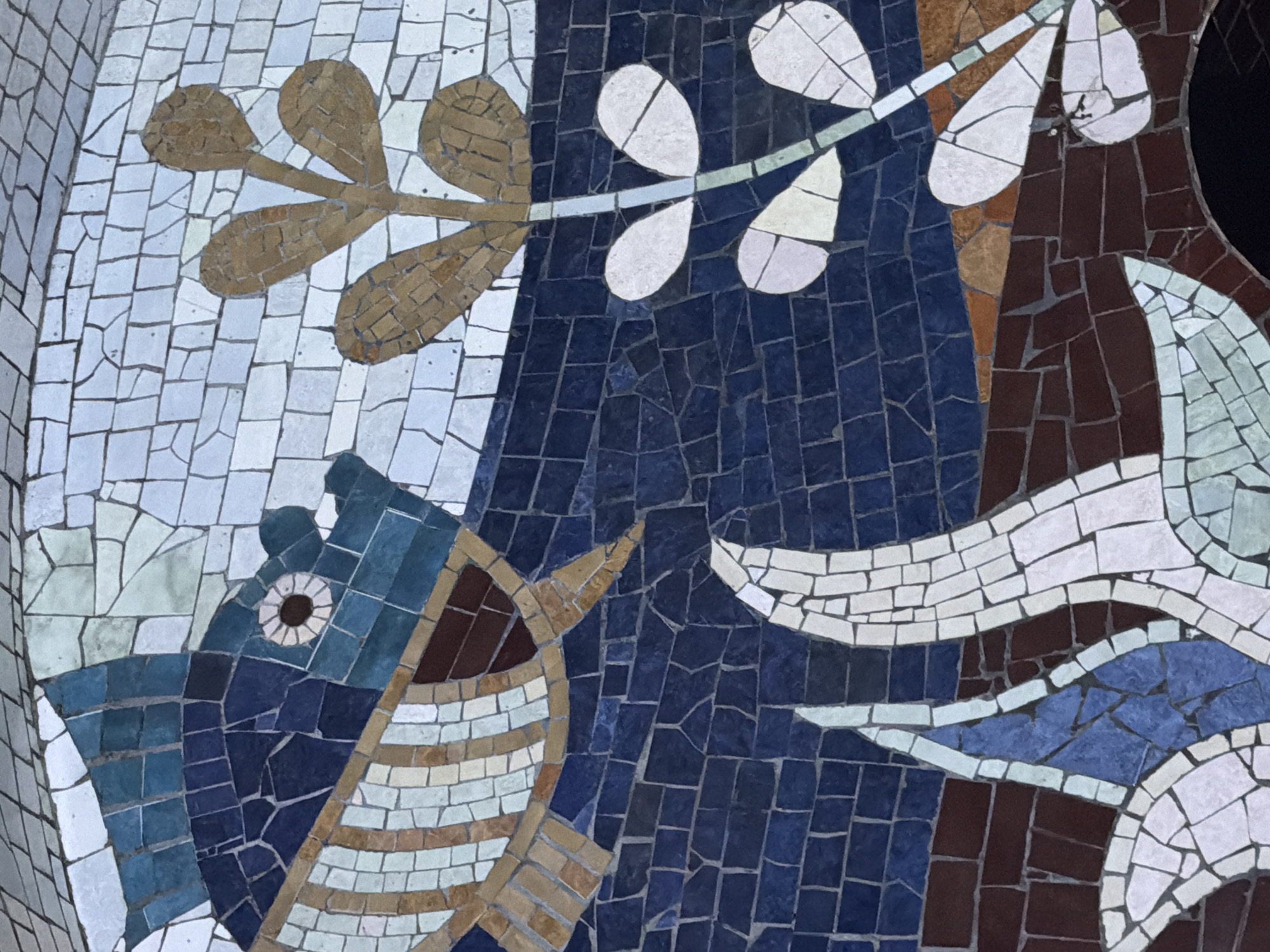 The image depicts a close-up of a mosaic mural in Tbilisi, Georgia. The mosaic consists of small, irregularly shaped tiles in various colors: blue, white, brown, and gold. The design incorporates stylized representations of a fish, leaves, and branches. The fish has a blue body with brown and white stripes. One branch is gold with rounded leaves, another white with rounded leaves. The background includes blue and white tiles. The mosaic appears to be part of a larger installation.