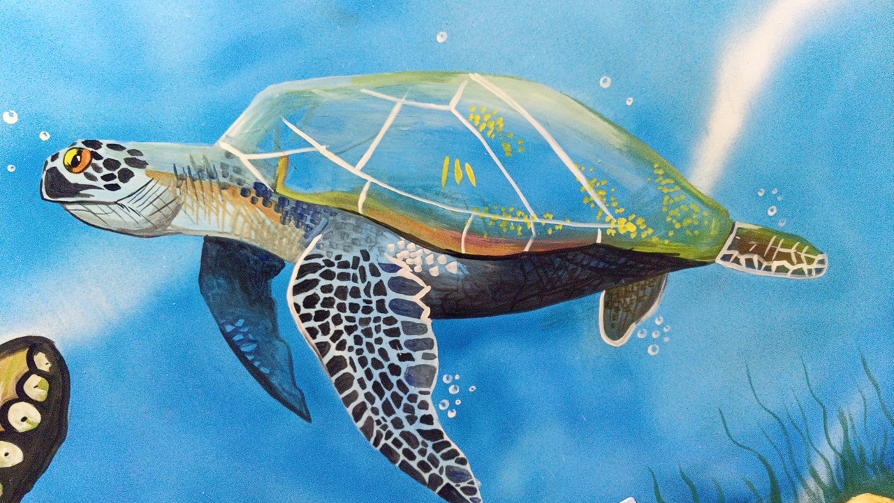 This is a close-up, eye-level, medium shot photograph of a mural depicting a sea turtle swimming in blue water. The sea turtle is painted in shades of green, yellow, and brown, with a patterned shell and flippers. There are bubbles rising around the turtle, suggesting it is underwater. The background is a gradient of blue, with some white streaks that could represent light or water currents. The mural is likely located on a wall in the city of Balykchy, Kyrgyzstan, given the context provided. The style of the painting is somewhat stylized and illustrative.