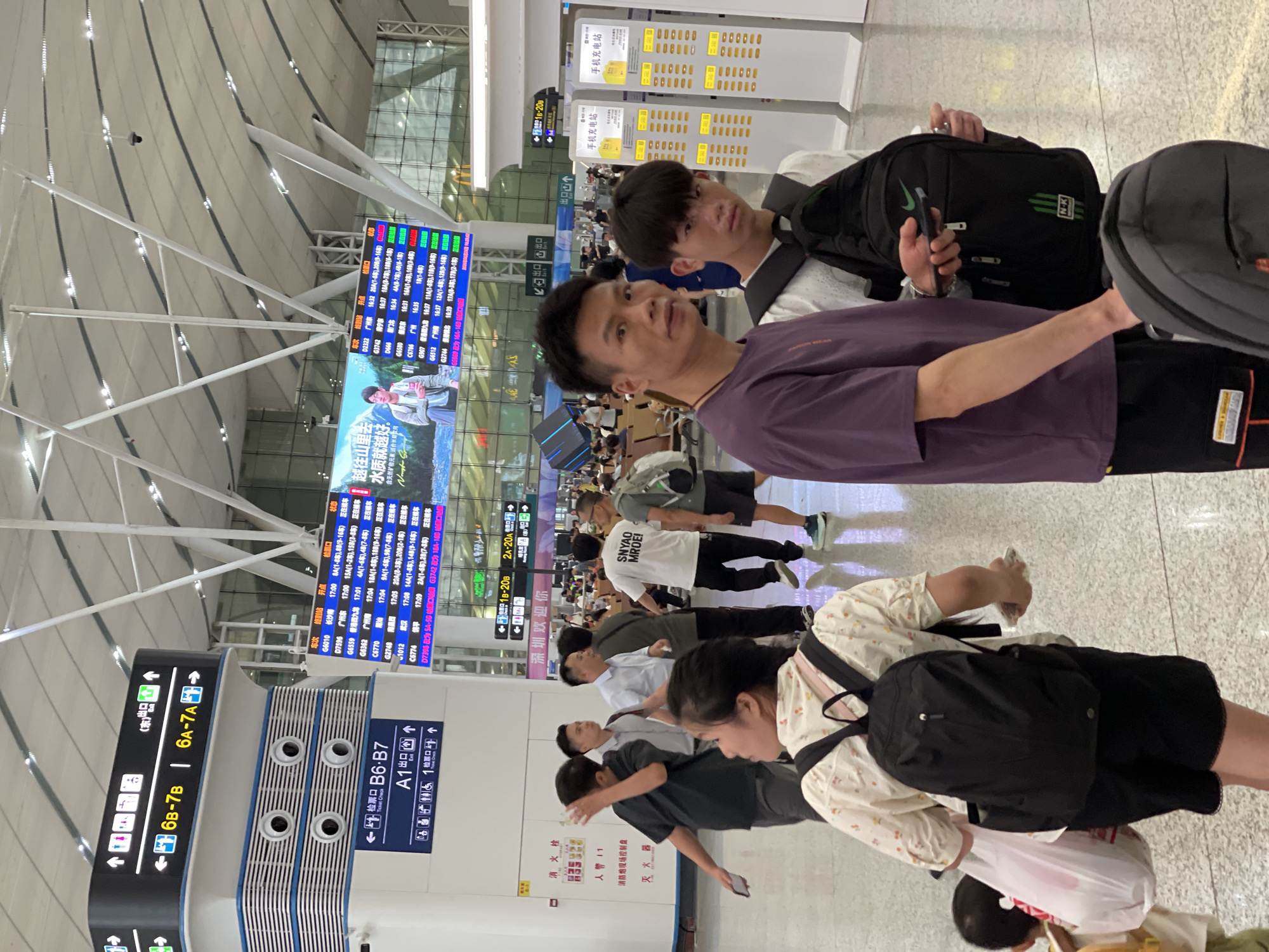 The image captures a scene inside what appears to be a busy transportation hub, possibly an airport or train station, given the departure boards and signage. The primary subjects are several individuals standing or walking, some with luggage, suggesting they are travelers. Two young men in the foreground are prominently featured, looking towards the camera. One is wearing a purple t-shirt, and the other a white t-shirt with text. Behind them, other travelers are visible, including people with backpacks and shopping bags.
The setting is indoors, characterized by a modern architectural style with a high ceiling and bright, artificial lighting. Multiple large digital display boards are visible, showing flight or train information, with columns listing departure times, destinations, and other relevant details. One board displays Chinese characters, including "越往山里去 水质就越好" which translates to "The further into the mountains, the better the water quality." Another sign reads "深圳欢迎你," meaning "Shenzhen Welcomes You." Charging stations for mobile phones are also visible, labeled "手机充电站" (mobile phone charging station).
The overall atmosphere appears to be one of transit and waiting. The individuals in the foreground seem to be posing for a photo, with the younger man in the white shirt smiling faintly. Other people in the background are more focused on their journeys, walking or standing in queues. The lighting suggests it could be daytime or evening, but the artificial illumination makes it difficult to determine the exact time of day. The presence of multiple departure boards and the general bustle indicate a public space with high foot traffic.