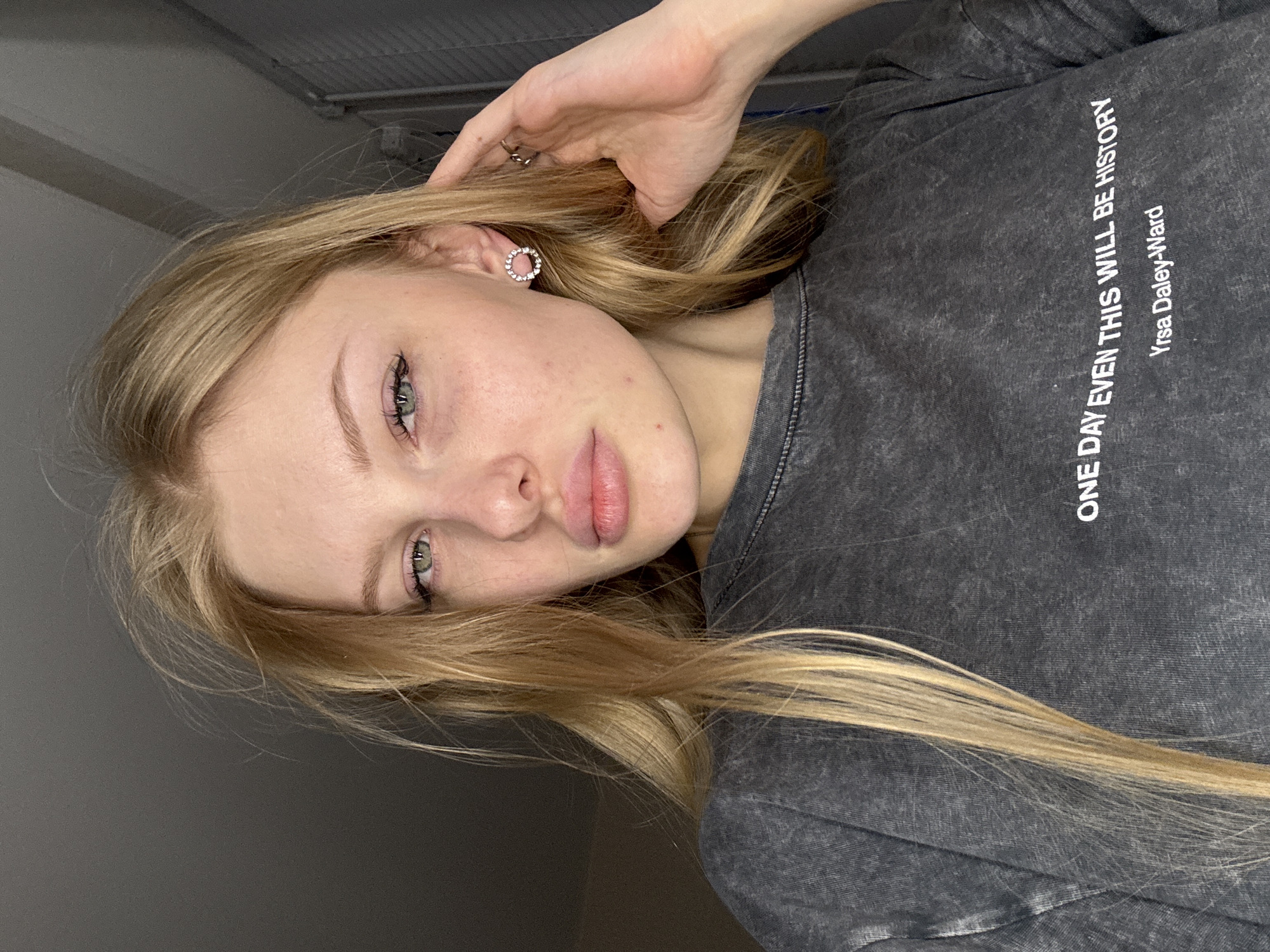 A light-skinned young woman with blonde hair and light-colored eyes looks at the camera. She is wearing an earring in her left ear and a gray long-sleeved shirt with the text "ONE DAY EVEN THIS WILL BE HISTORY Yrsa Daley-Ward" printed in white. She is holding her hair with her left hand. The background is gray. The location is not specified in the image.