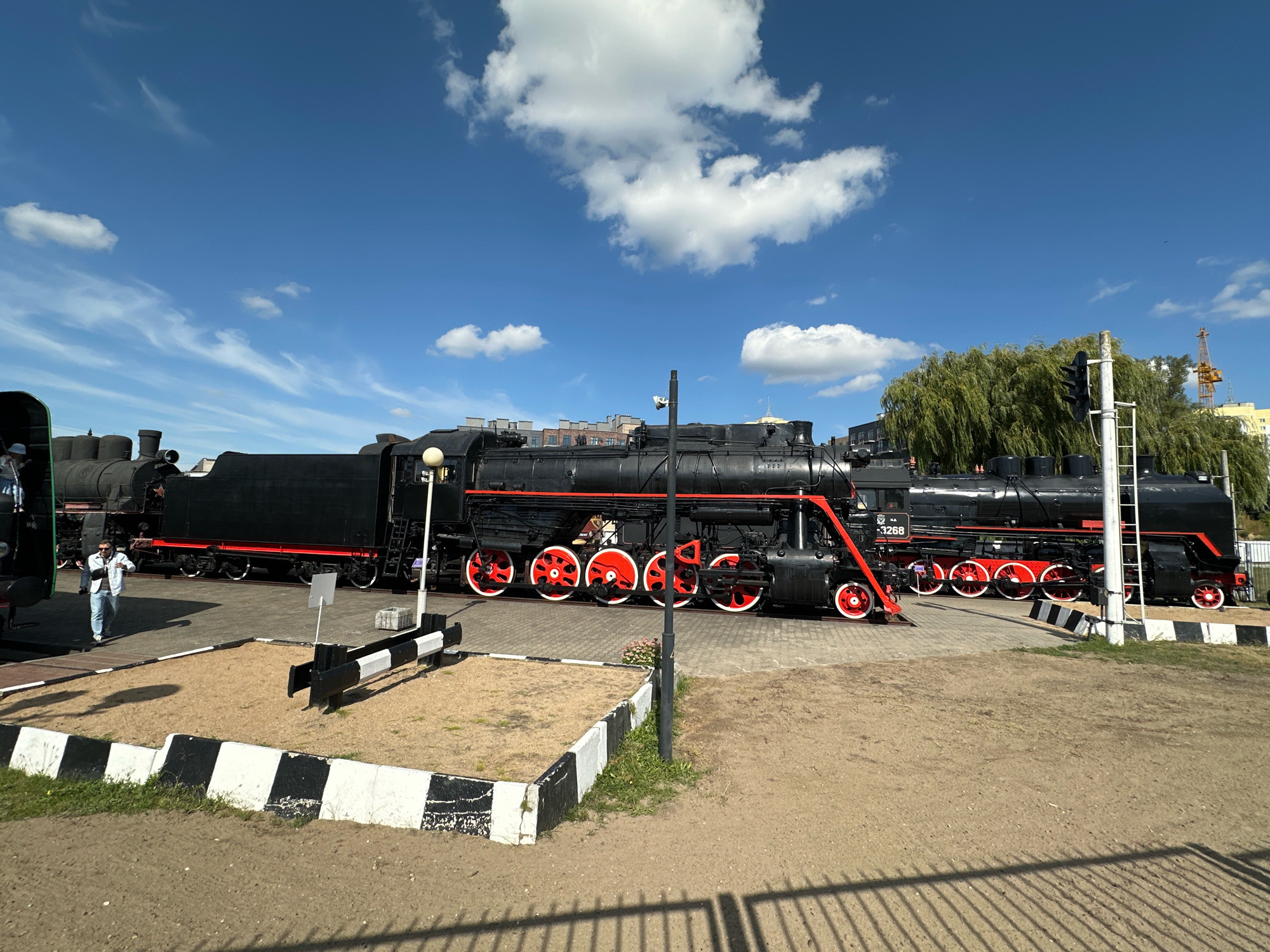 In Brest, Belarus, a bright, sunny day is captured with a clear blue sky dotted with fluffy white clouds. The scene showcases a row of large, black steam locomotives parked on a railway track. The locomotives are adorned with red accents on their wheels and along their bodies. In the foreground, a person wearing a light-colored jacket and blue jeans walks away from the viewer towards the locomotives. The ground is a mix of sand and pavement, with a black and white striped border running along a sandy area. A traffic light and a tall, green willow tree are visible in the background. A construction crane is also present in the distance, suggesting ongoing development in the area.