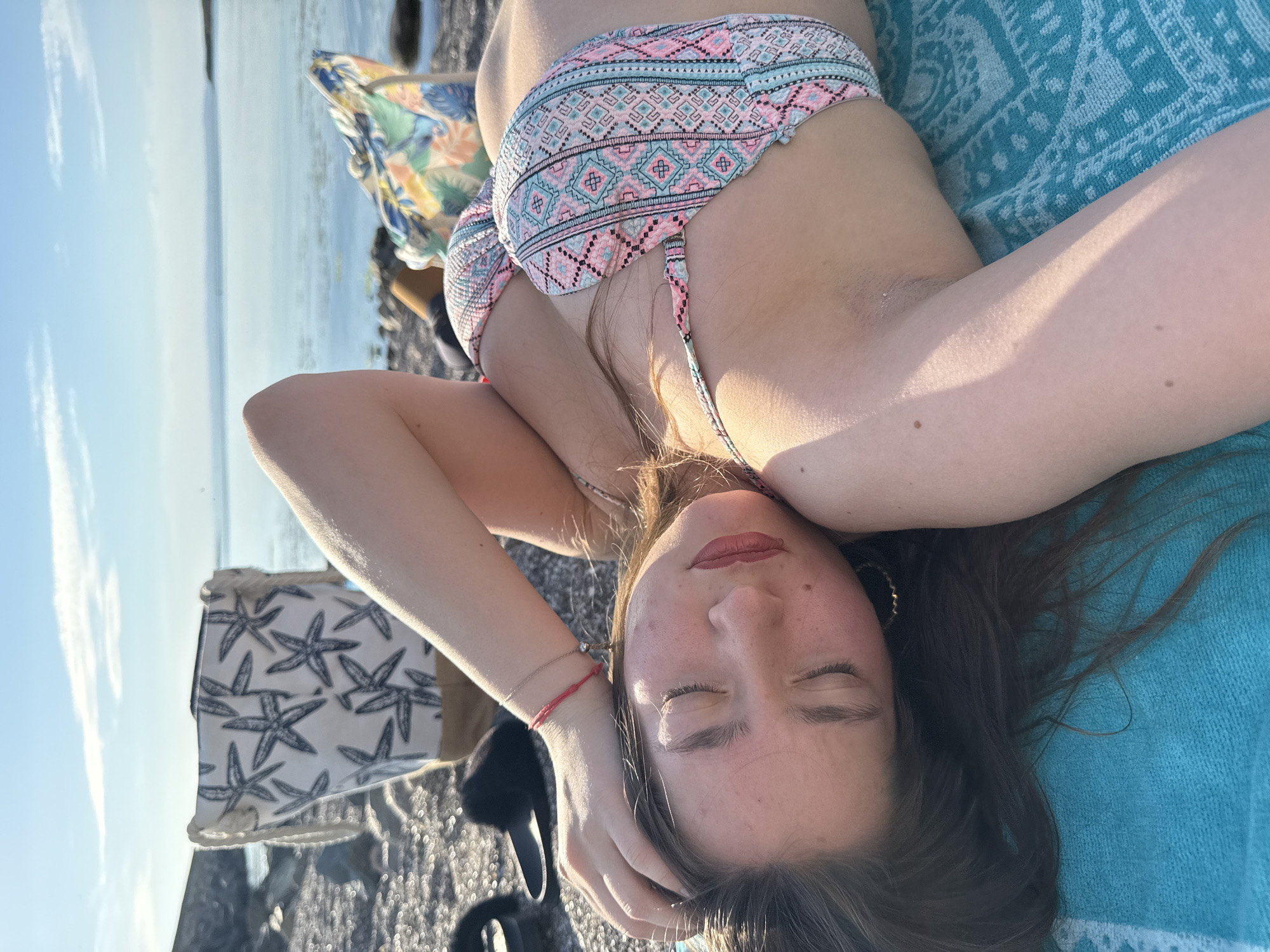 A young woman lies on a blue towel on a pebble beach in Sozopol, Bulgaria. She is wearing a patterned bikini and her eyes are closed, suggesting she is relaxing or sunbathing. Her head is resting on her arm, and her long, dark hair is spread out. A colorful patterned fabric, likely a beach bag or umbrella, is visible behind her. The sea is in the background, with a calm surface reflecting the clear sky and some clouds. The lighting suggests it is either early morning or late afternoon, with the sun casting long shadows. The scene conveys a sense of peace and enjoyment of a summer day by the sea. There is no visible text in the image.