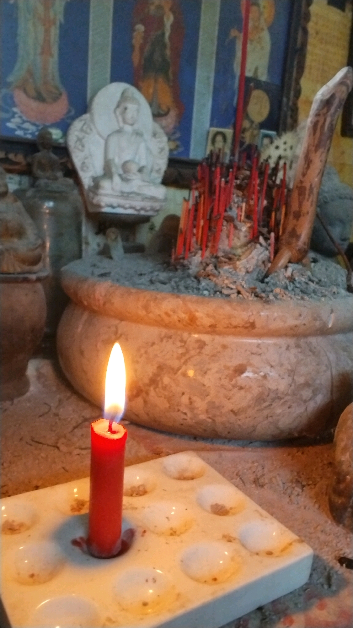 This is a close-up, medium shot of an altar, likely in a home or shrine, in Penompo, Indonesia. In the foreground, a lit red candle stands in a white, multi-compartment holder. The flame is bright and illuminates the surrounding area. Behind the candle, a large, round, stone-like pot is filled with ash and numerous red incense sticks, some of which appear to be burning or recently extinguished. The pot is positioned on a sandy surface. In the background, there is a small white statue of a seated figure, possibly a Buddha or a deity, with a halo. Behind the statue, a dark blue tapestry or painting with figures is visible. To the right of the incense pot, a piece of weathered wood protrudes upwards. The overall atmosphere is one of reverence and ritual.