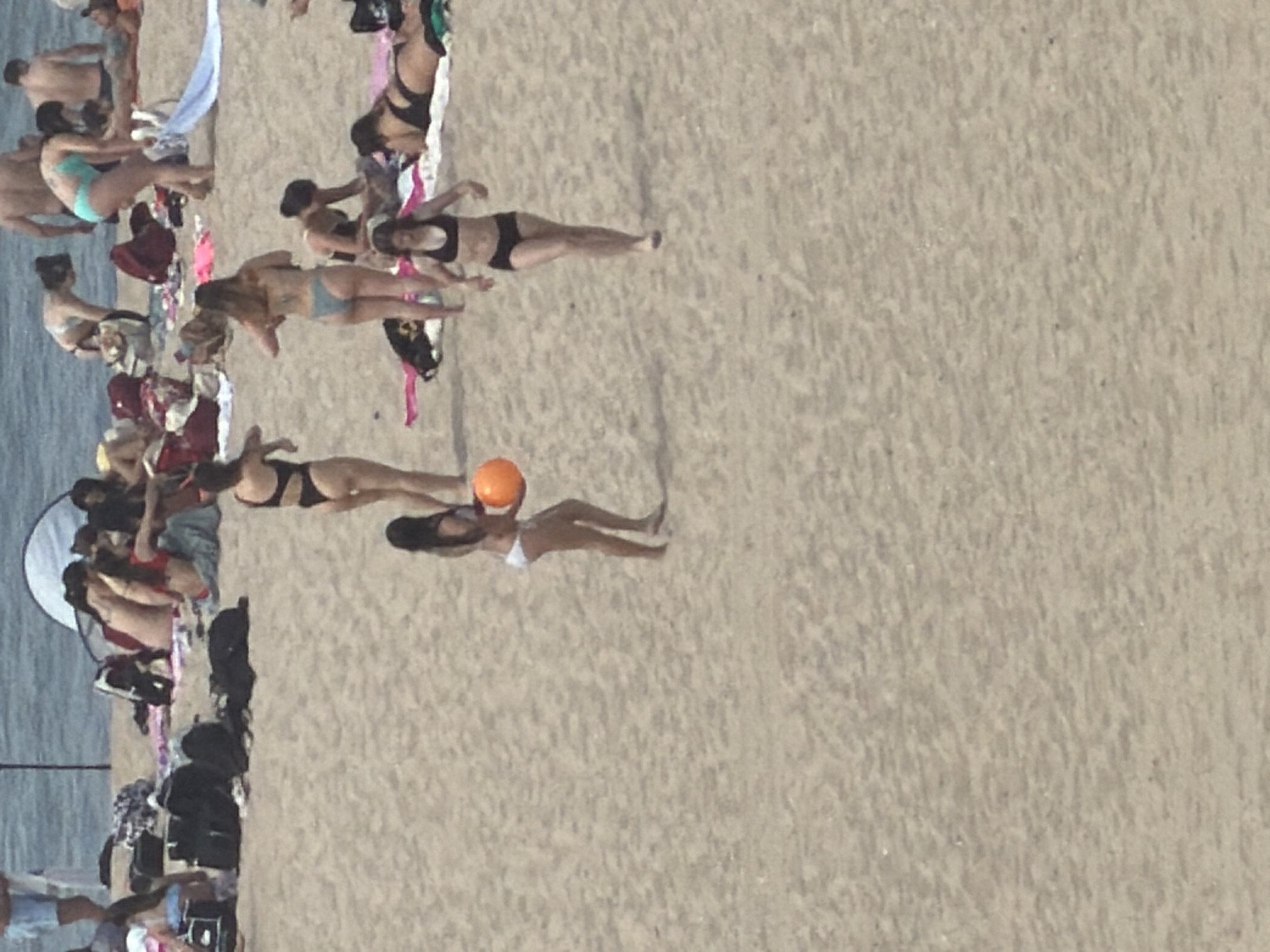 This aerial, full-frame photograph depicts a crowded beach scene at Praia da Bafureira in Espinho, Portugal. The sandy beach is populated with numerous individuals, many of whom are wearing swimwear. In the foreground, a woman in a black bikini is walking towards the viewer, her body angled to the right. Behind her, another woman, also in a black bikini, is walking from right to left and holding an orange ball. Further in the background, groups of people are scattered across the sand, some sunbathing, others standing or walking. Beach umbrellas and towels are visible, along with several black bags and backpacks. The ocean is visible in the top portion of the image, with a large, white tent-like structure situated on the beach near the water's edge. The overall scene conveys a typical summer day at a popular beach.