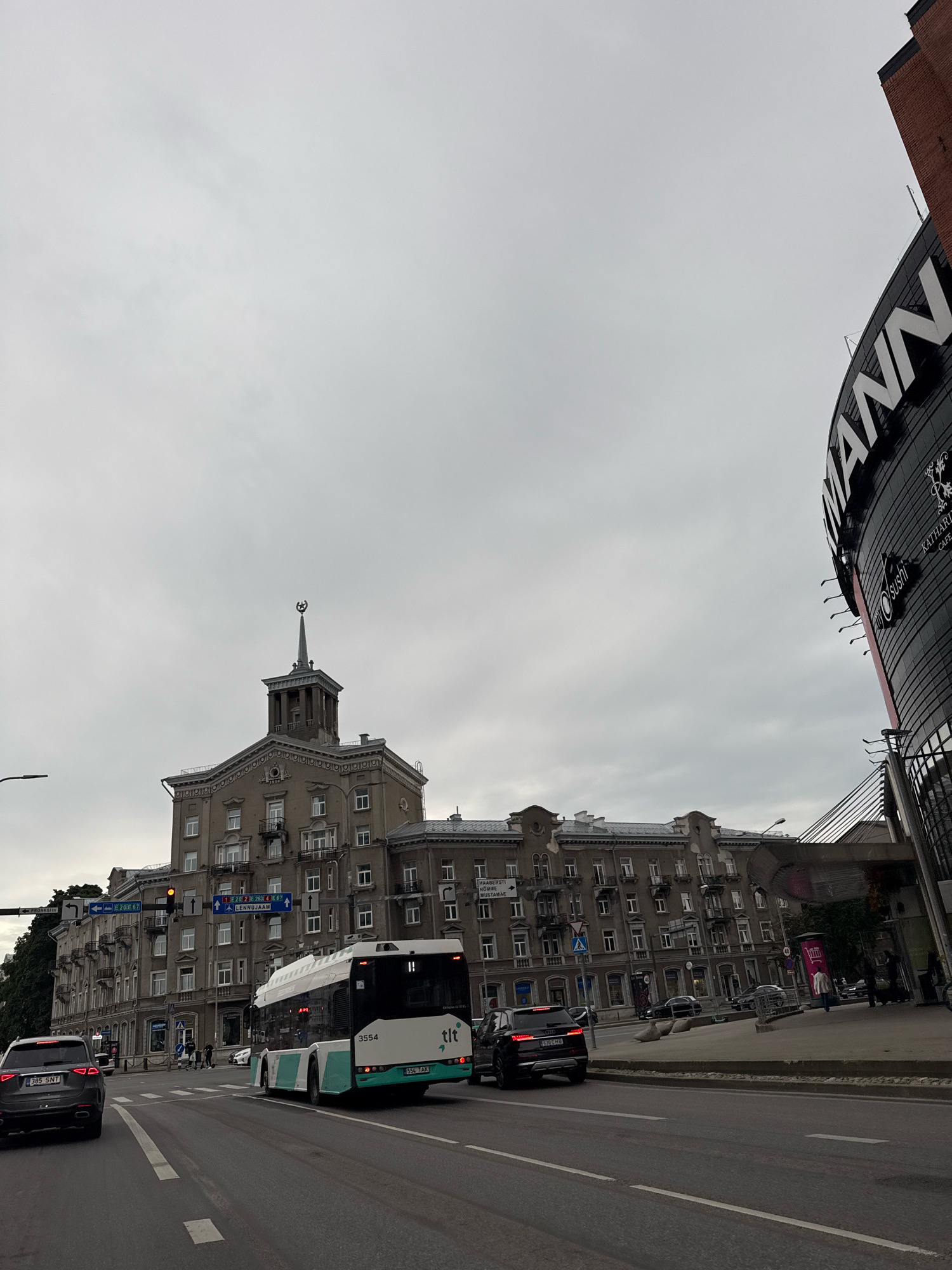 The image captures a street scene in Tallinn, Estonia, with a focus on the intersection of Narva Road and Pronksi Street. In the foreground, a dark grey Audi Q7 SUV with license plate "385 SNT" is visible on the right lane of the road. To its left, a white and teal bus, marked with "3554" and "tlt" (Tallinna Linnatransport), is turning across the intersection.
The background features a large, ornate building with a distinctive spire topped with a star-like emblem, a common architectural style from the Soviet era. To the right of this building, a modern, curved structure with a prominent "MANN" logo is visible, likely a commercial building or shopping center.
The sky is overcast, suggesting a cloudy day. Pedestrians can be seen crossing the street near the bus. Traffic lights are red for the direction of the Audi and the bus. Road signs in Estonian indicate directions to various locations, including "PIRITA," "LASNAMÄE," and "LENNUKAJAM." The overall impression is that of a busy urban intersection under a gray sky.