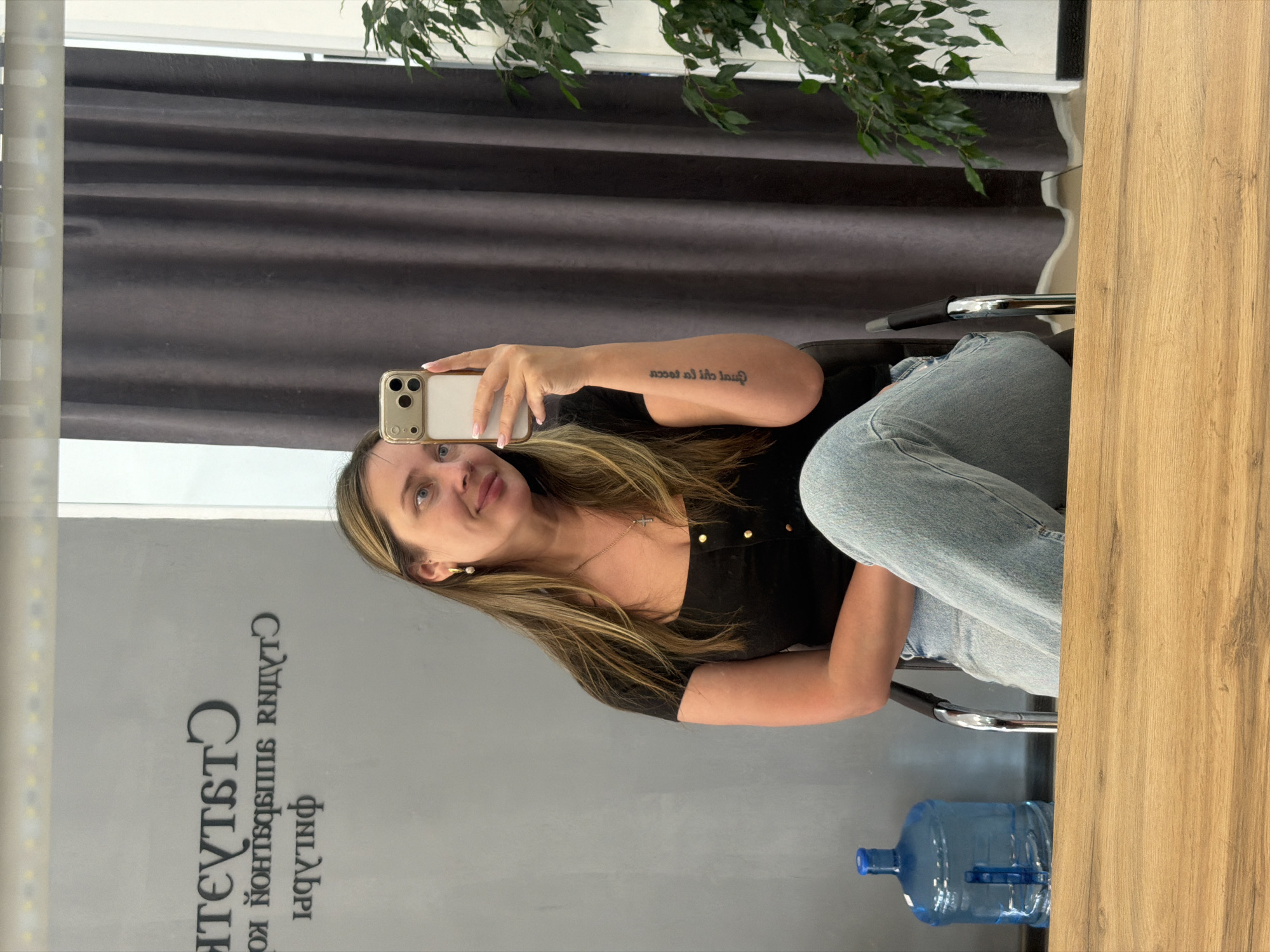 A woman is captured in a mirror selfie, seen from the waist up, smiling slightly as she holds a light-colored smartphone. She has long, straight brown hair with lighter highlights and is wearing a black collared top with gold buttons, paired with light blue jeans. A delicate silver cross necklace hangs around her neck, and she wears a small pearl earring in her visible ear. On her right forearm, a tattoo in Cyrillic script reads, "послал ей на счастье," which translates to "sent to her for happiness."
The setting appears to be an indoor commercial space, possibly a studio or salon, located in Chaikovsky, Russia. Behind the woman, dark gray pleated curtains or blinds are visible. To her left, a leafy green plant adds a touch of natural decor. The mirror reflects text, which, when read correctly, includes Cyrillic words such as "СТУДИЯ" (STUDIO) and "ФИЛАБРИ" (FILABRI), suggesting the name of the establishment. A large, clear blue water bottle with a handle rests on the floor to her right, partially obscured by a wooden panel or counter. The overall impression is casual and well-lit.