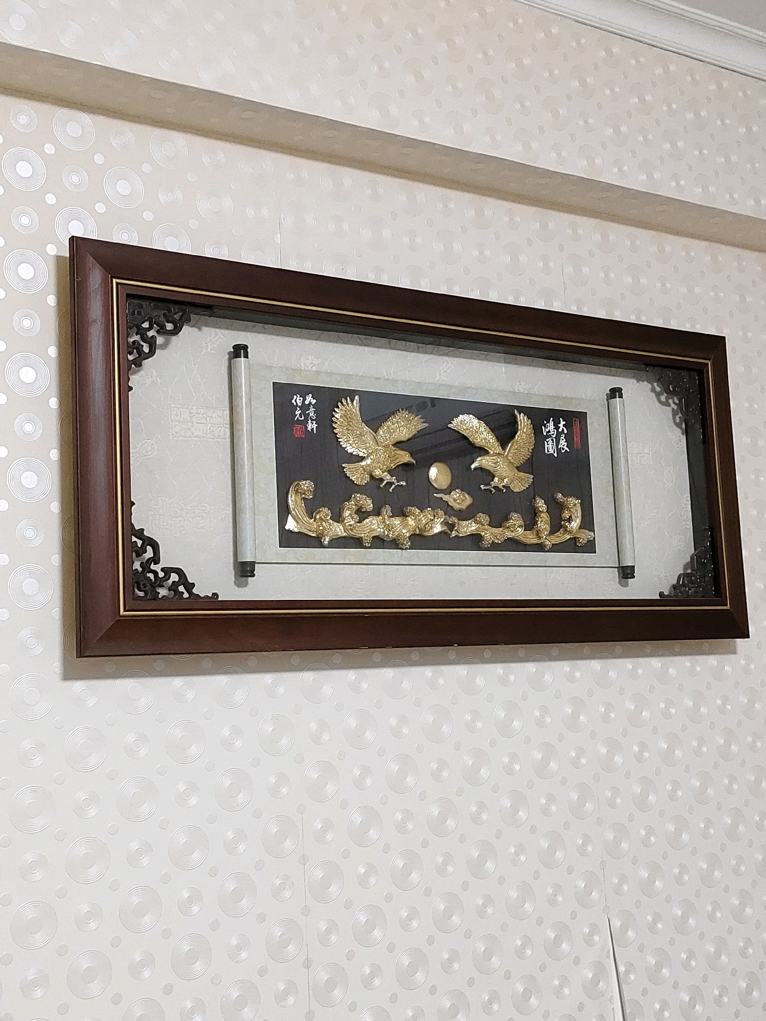 An ornate, framed artwork hangs on a light-colored wall with a circular pattern. The artwork is a rectangular piece with a dark brown wooden frame. Inside the frame, there is a scroll-like element on the right and a dark background on the left. The dark background features two golden eagles, a golden sun or moon, golden clouds, and a golden dragon or serpentine creature at the bottom. There are also red and black Chinese characters on the dark background. The wall has a raised, concentric circle pattern. The scene is indoors, and the lighting appears to be artificial. There are no people visible in the image. The artwork is positioned horizontally, centered within the frame, and appears to be a decorative piece for a wall. The location is not discernible from the image itself.