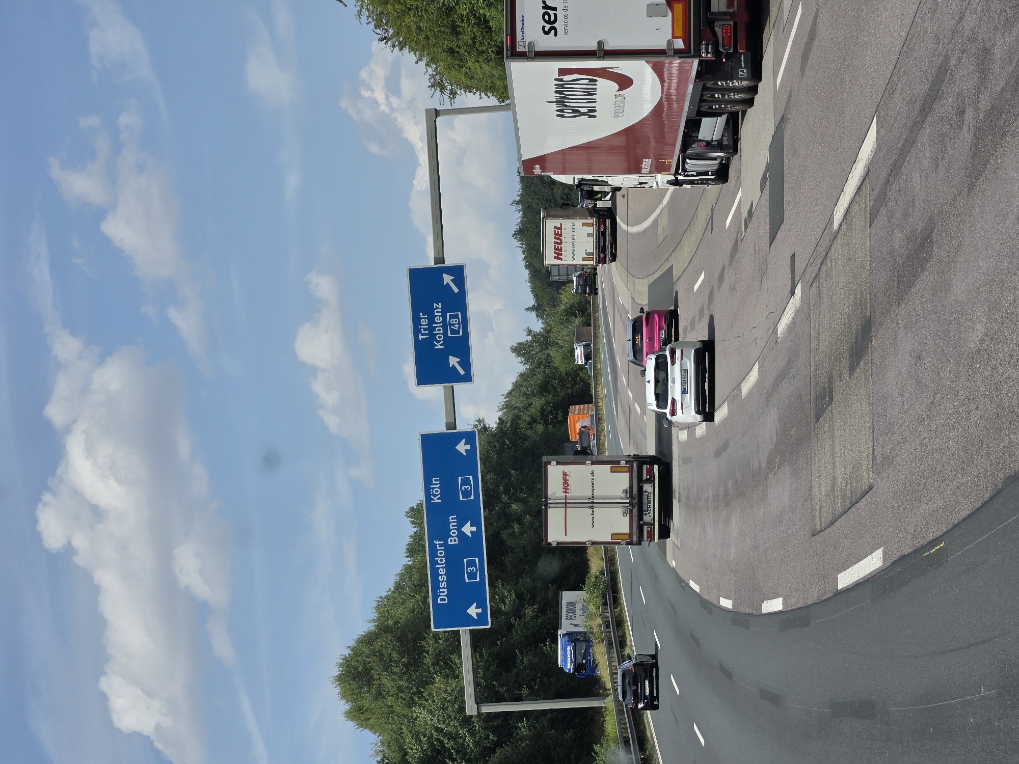 The image shows a multi-lane highway with traffic. Overhead signs indicate directions to Düsseldorf, Bonn, and Köln (Cologne), and Trier and Koblenz. Several trucks and cars are visible on the road. The environment is characterized by a blue sky with white clouds and dense green trees lining the sides of the highway. The weather appears to be clear and sunny. This scene is likely a portion of the A3 autobahn near Dernbach (Westerwald), Germany, a known junction for these routes.