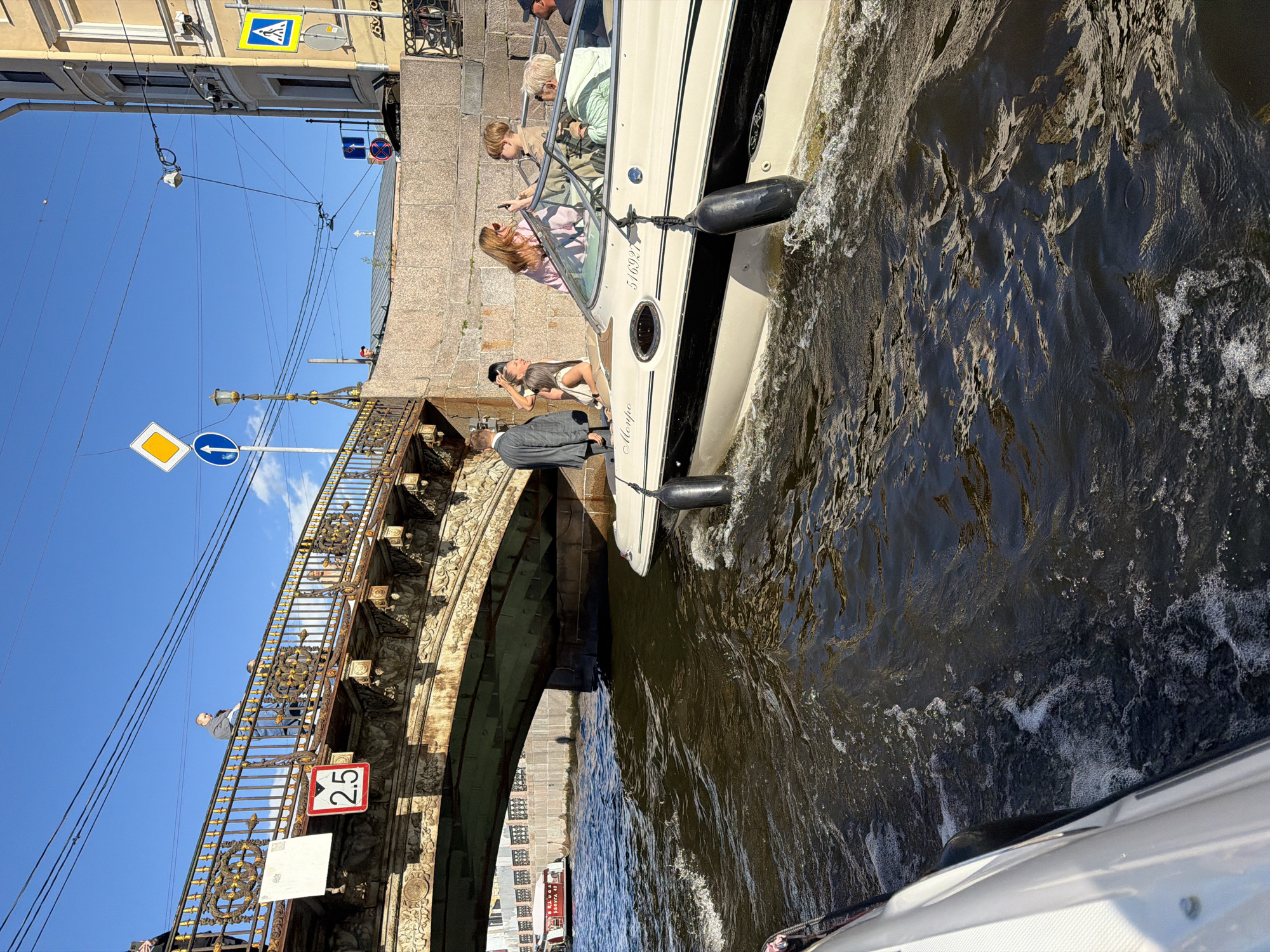 The image shows a sunny day in Saint Petersburg, Russia, with a view from a boat on a canal. In the foreground, a white boat is moving through dark, choppy water, creating a wake. Another boat, longer and with a white canopy, is visible under a bridge in the background, bearing the Cyrillic text "СЕВЕРНАЯ 42".
The bridge, an ornate metal structure with golden railings and decorative elements, spans the canal. On the bridge, several people are standing and observing the surroundings. One individual in a grey suit stands on the left, while a group of three women and a child are on the right. One woman, with long red hair, is holding up a phone, seemingly taking a photo or video. A young boy next to her is also looking at the phone. Another woman with blonde hair is also present, and an older woman with short grey hair is seated.
Further in the background, a stone embankment lines the canal, with buildings visible beyond. Traffic signs, including a yellow diamond-shaped sign with a blue arrow and a pedestrian crossing sign, are visible on poles near the bridge. The sky is a clear blue with a few wispy clouds. The overall scene captures a leisurely boat trip on a picturesque canal in Saint Petersburg.
