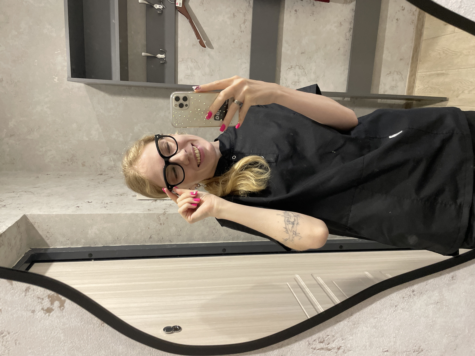 A young, fair-skinned woman with blond hair tied back in a partial ponytail, wearing black-framed glasses, a black top, and pink nail polish, is taking a selfie with her smartphone. The phone's case is clear with glitter-like dots. She's smiling and holding the phone up in her right hand. Her left hand is also raised near her face. There is a tattoo on her left forearm. The background consists of a grey and white patterned wall with a wall-mounted shelf containing hooks and a hanger. This photo was taken in the city of Izhevsk, Russia. The image is a mirror reflection.